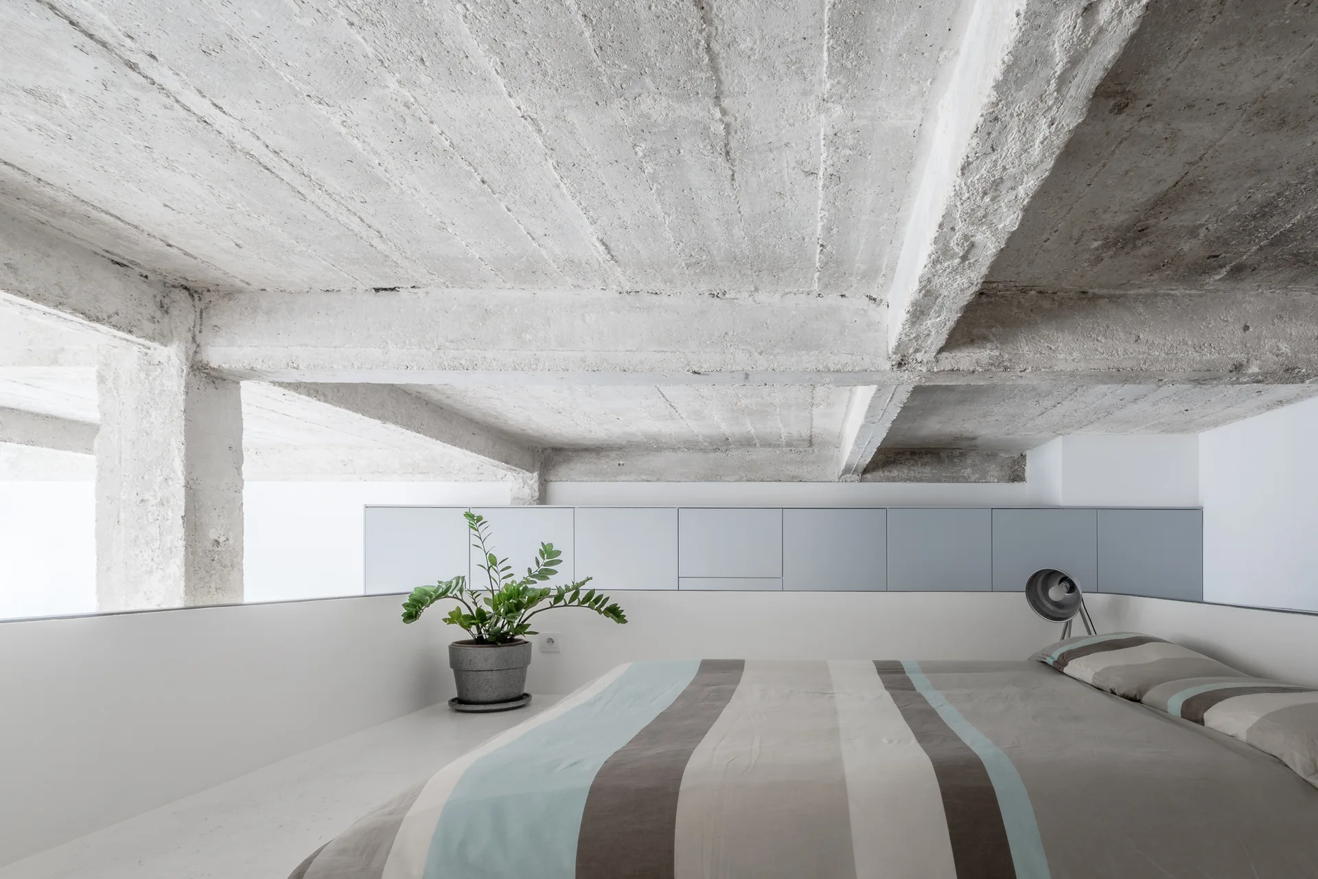 A minimalist loft bedroom features a bed with striped bedding beneath a rough, exposed concrete beam ceiling and a row of light grey cabinets.