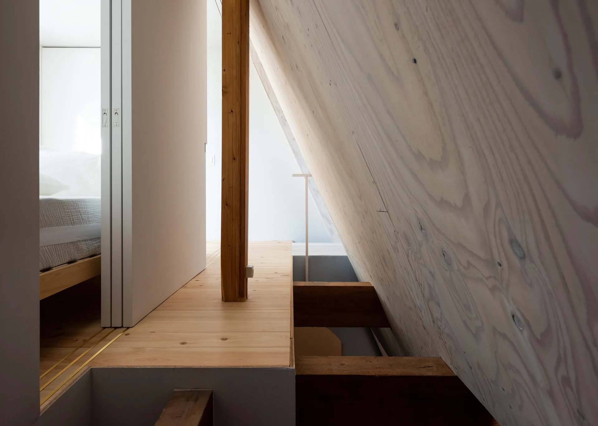 A minimalist attic loft bedroom features a sloped plywood wall, exposed wooden beams, and a sliding white door revealing a simple bed.