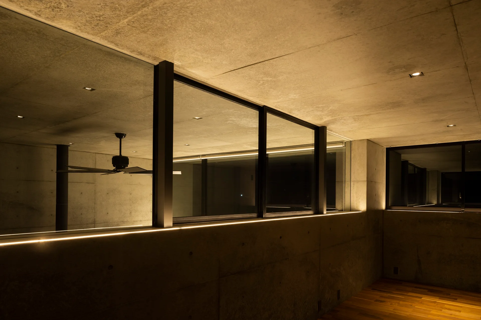 An industrial concrete interior featuring black-framed glass windows, a warm linear cove light, and a glimpse of hardwood flooring.