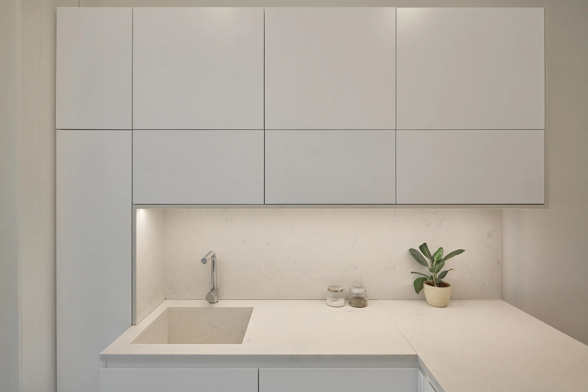 A minimalist kitchen features white handleless cabinets, a stone-like countertop and backsplash, an integrated sink, and warm under-cabinet lighting.
