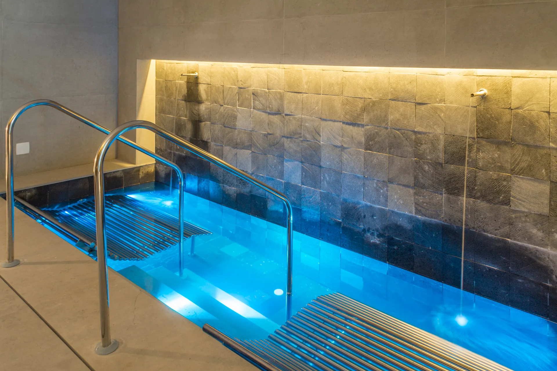 An indoor spa pool features illuminated blue water, stainless steel handrails, and a textured grey stone tile wall highlighted by warm recessed lighting.