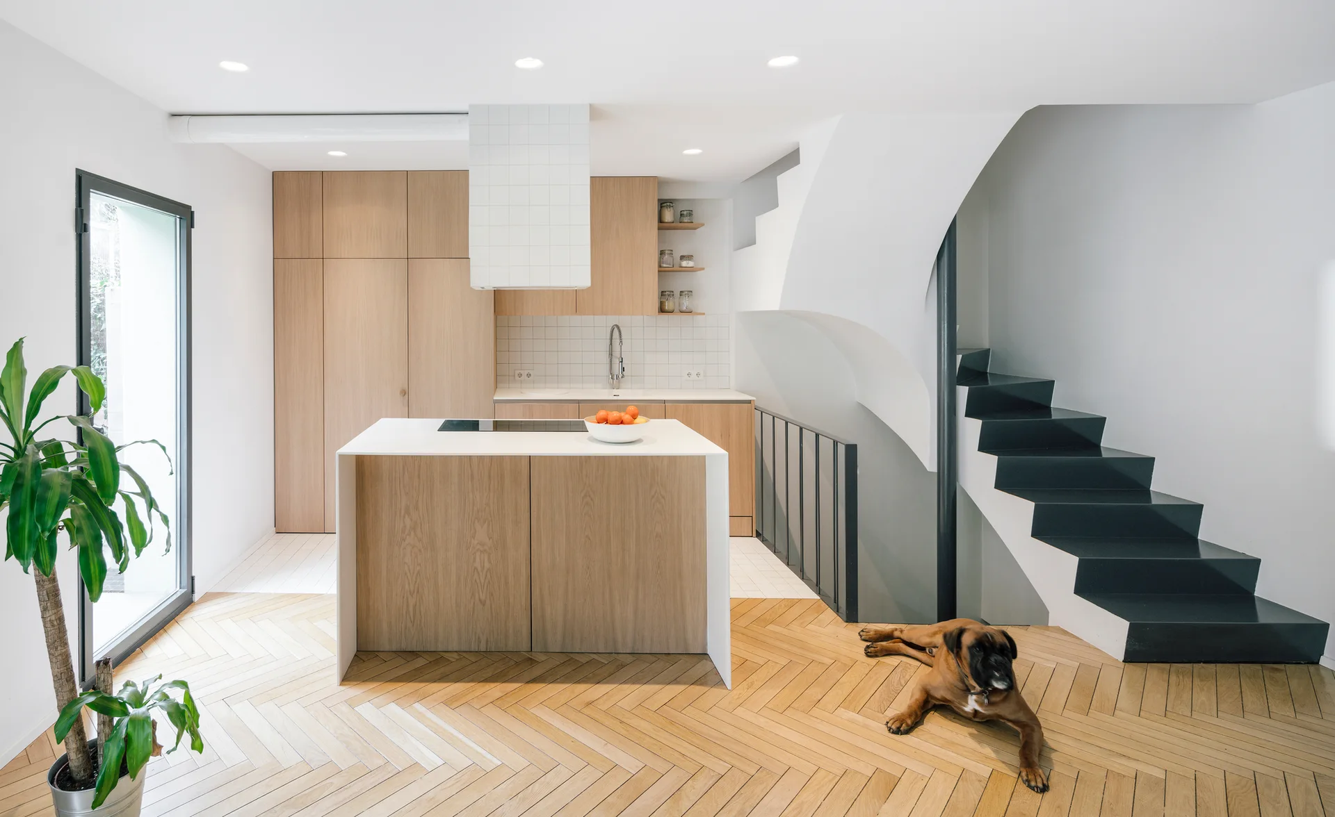 A minimalist kitchen features light wood cabinets, a matching island, herringbone floors, and an adjacent black metal staircase.
