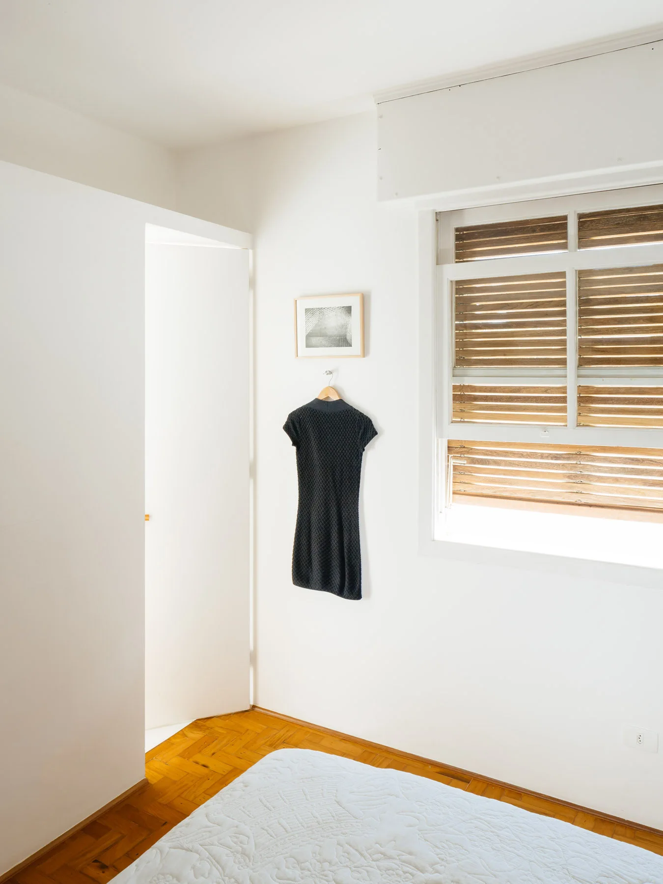 A minimalist white bedroom featuring natural wood parquet flooring, horizontal wooden window blinds, and a black garment hung below a small framed picture.
