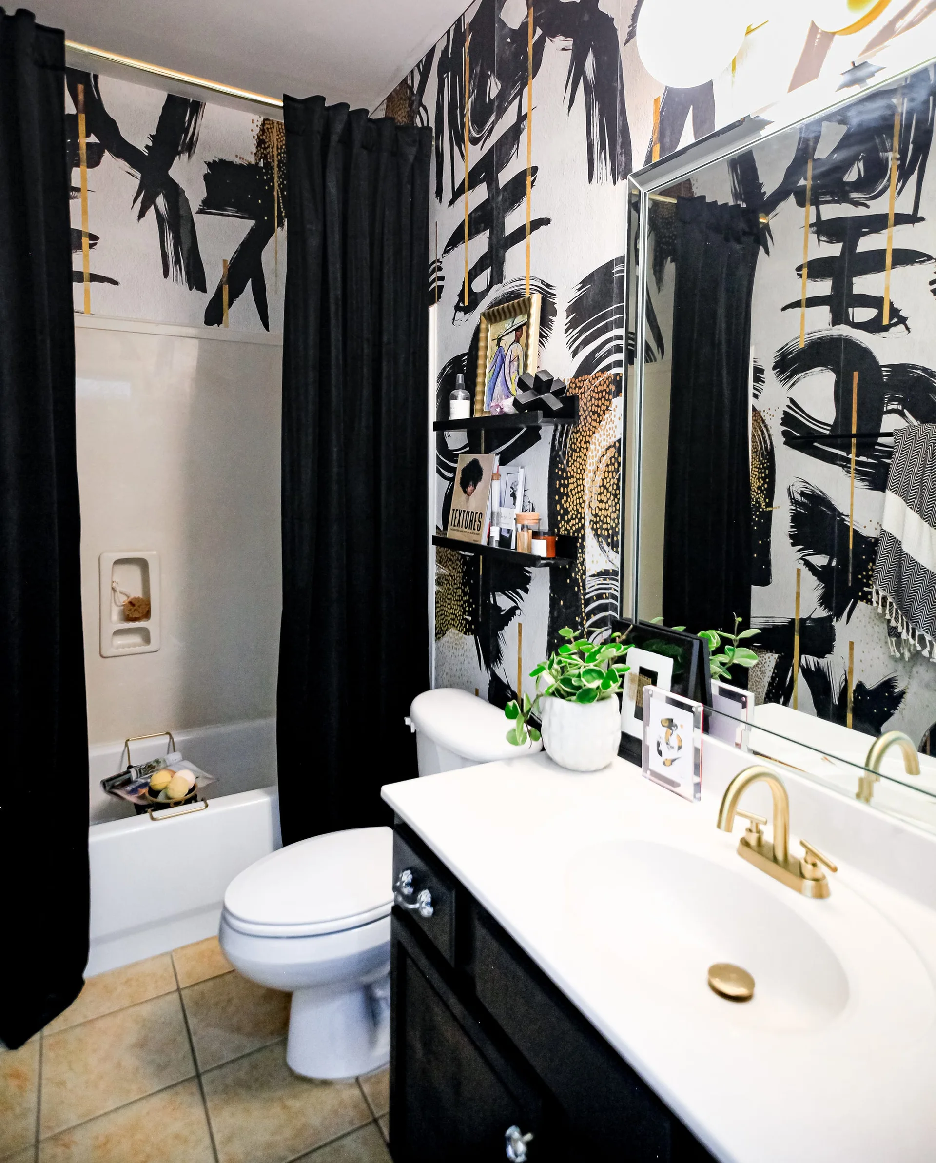 A bathroom features abstract black and white graphic wallpaper, a black vanity with a gold faucet, a white toilet, and black shower curtains.