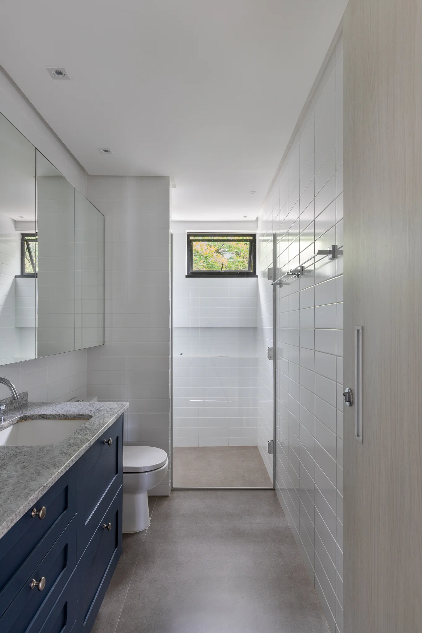 A narrow contemporary bathroom with a navy blue vanity, white subway tile walls, and a walk-in shower in the background.