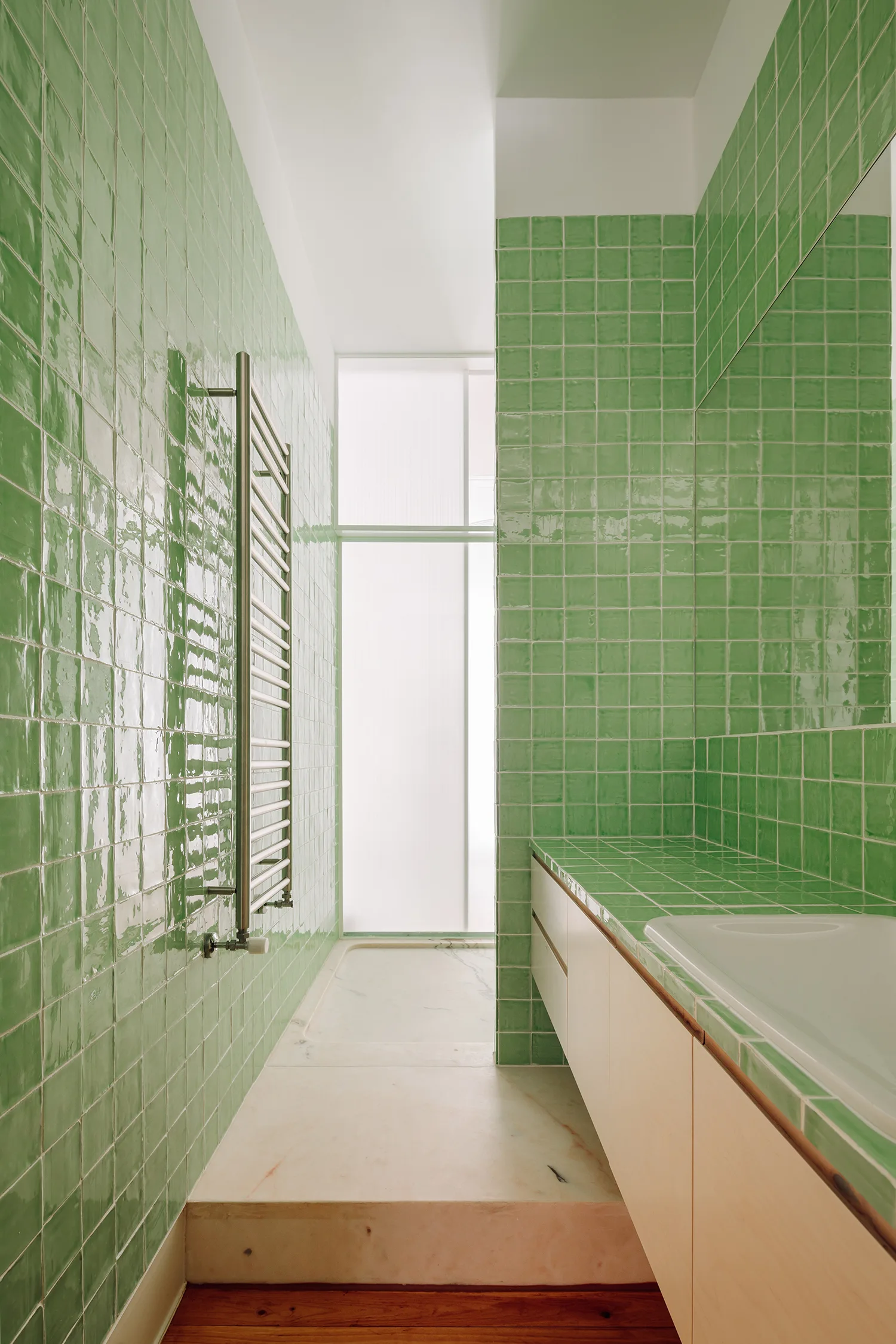 A narrow contemporary bathroom features glossy green square tiled walls, light wood cabinetry, a built-in bathtub, and a metal towel warmer.