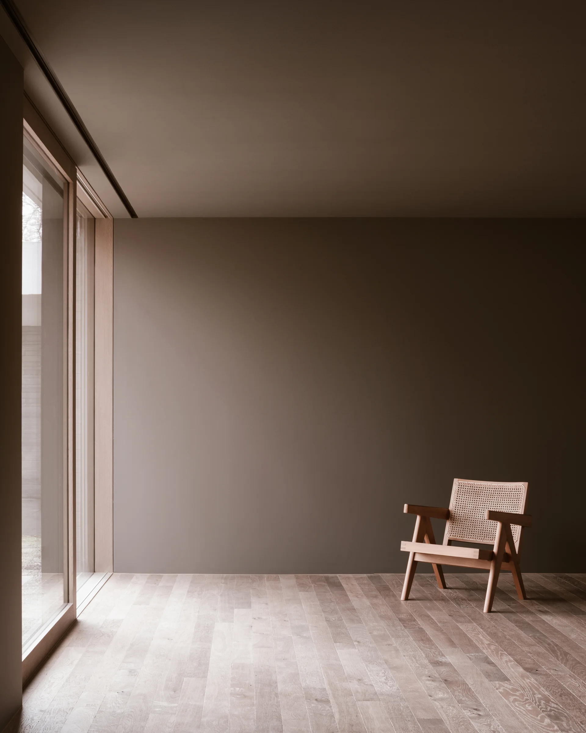 A minimalist room with solid taupe walls and light wood floors features a single wooden armchair with a cane back next to a large glass door.