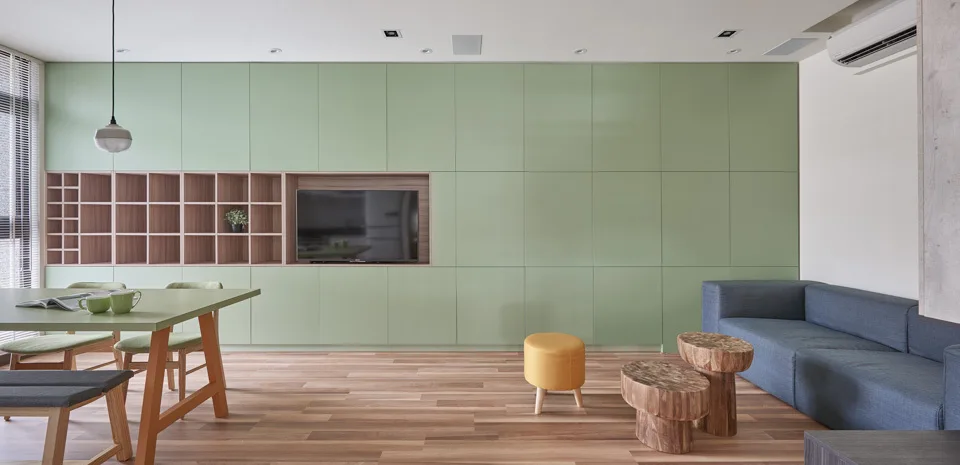 A modern living and dining space featuring a custom sage green storage wall with integrated wood shelving, a blue sofa, and wood flooring.