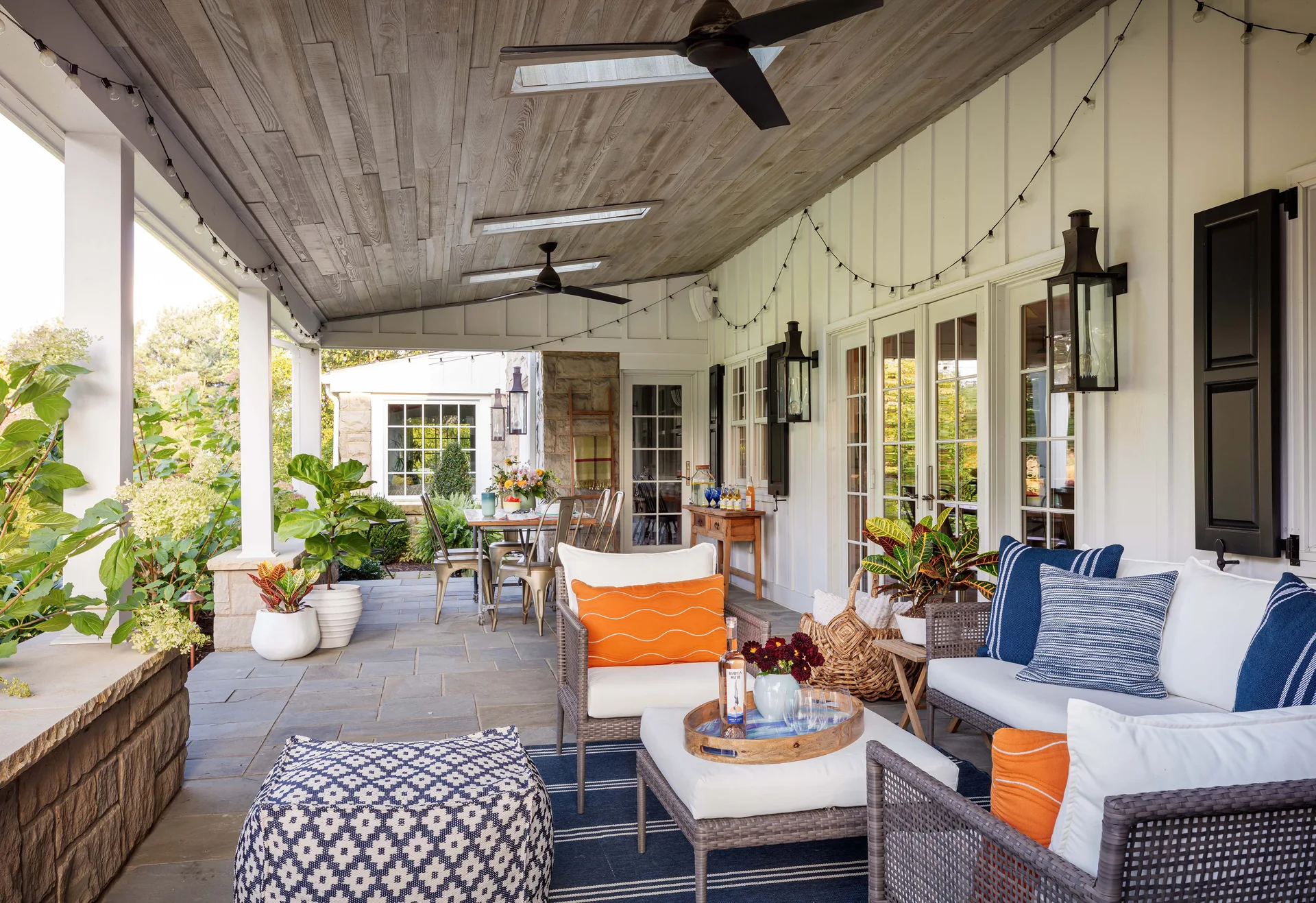 A covered outdoor patio featuring grey stone flooring, a wood plank ceiling, and wicker seating with blue and orange accent pillows.