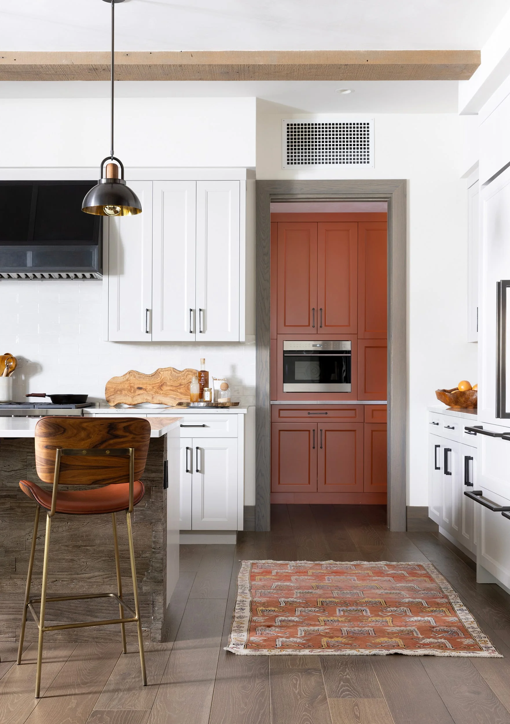 A bright kitchen features a wooden-detailed island, a vintage-style runner rug, and a doorway looking into a built-in rust-red pantry.
