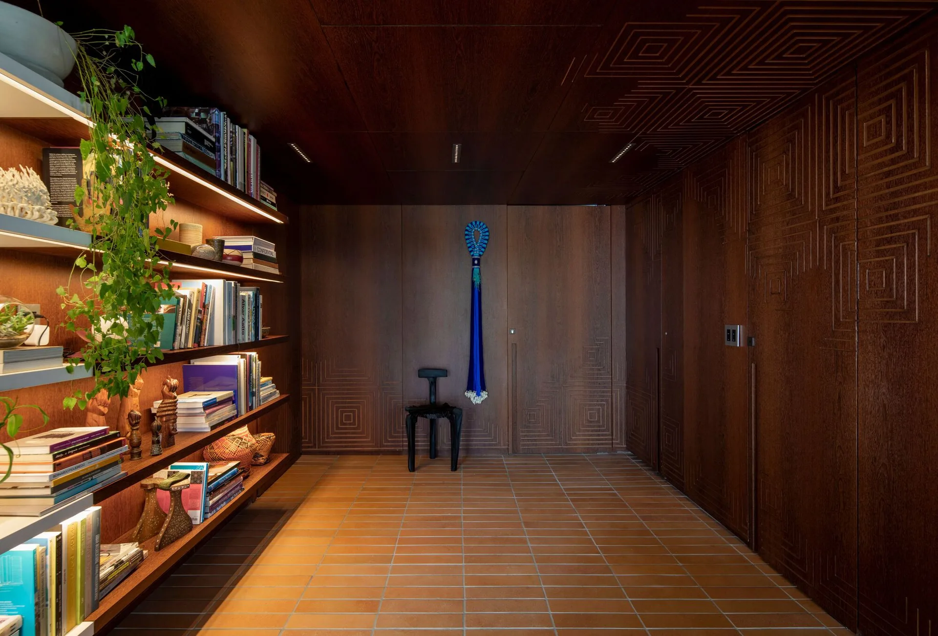 A library features carved dark wood paneling on the walls and ceiling, built-in illuminated shelving with books, and terra-cotta tile flooring.