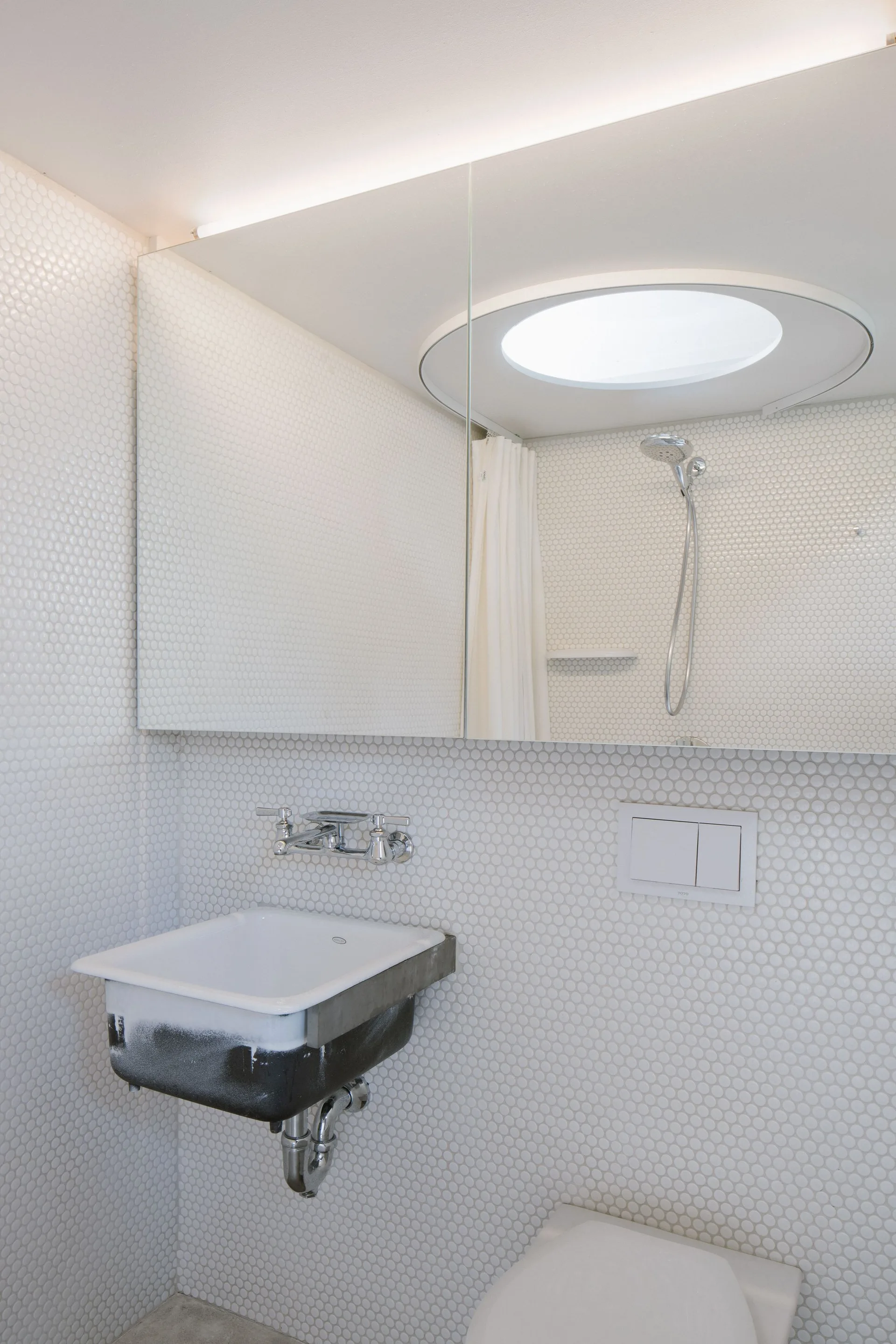 A white penny-tiled bathroom featuring a wall-mounted sink with chrome fixtures and a large mirror reflecting a skylight and shower.
