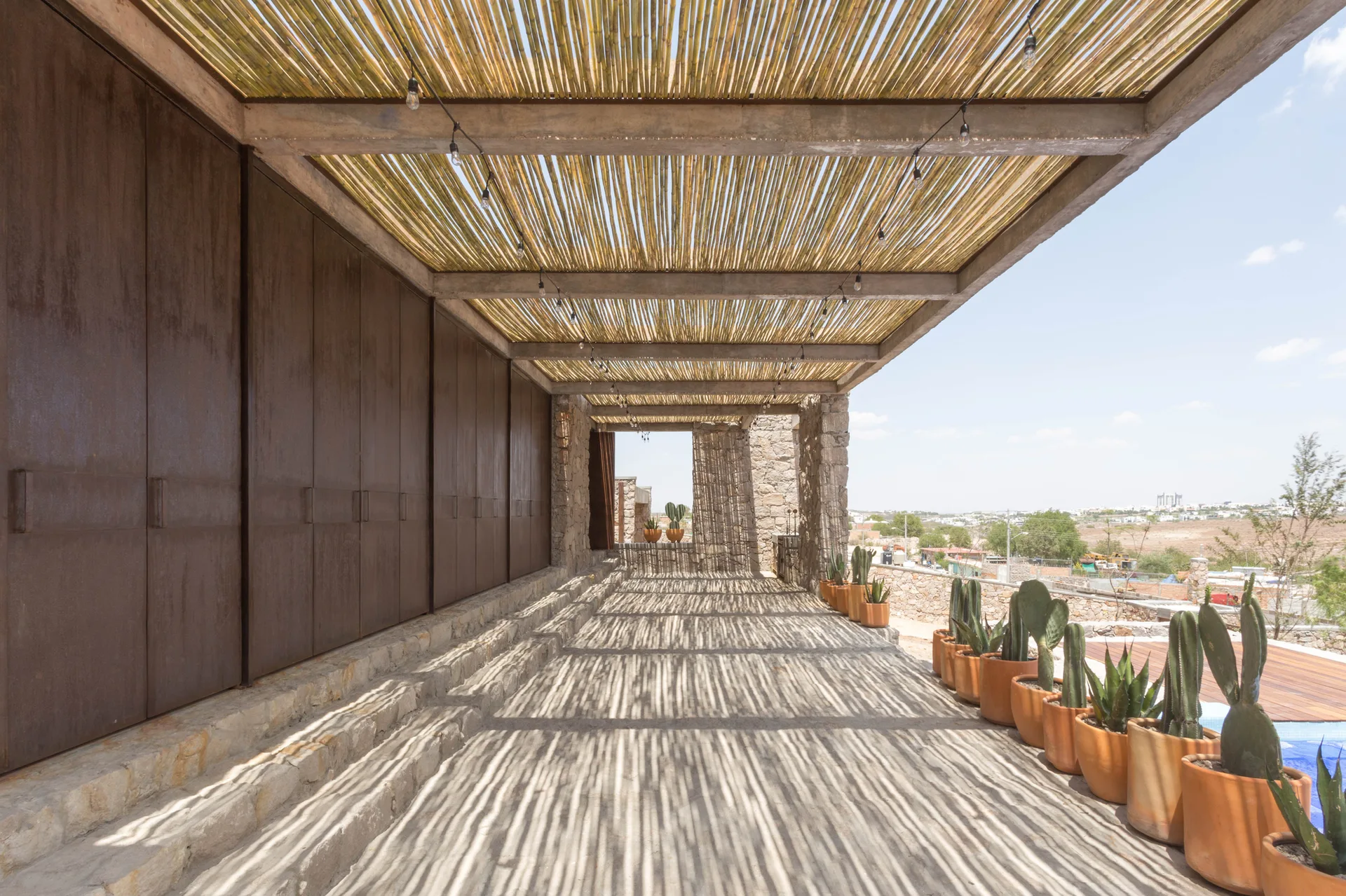 A covered outdoor patio featuring a slatted bamboo pergola roof casting linear shadows onto a stone floor alongside a row of potted cacti.