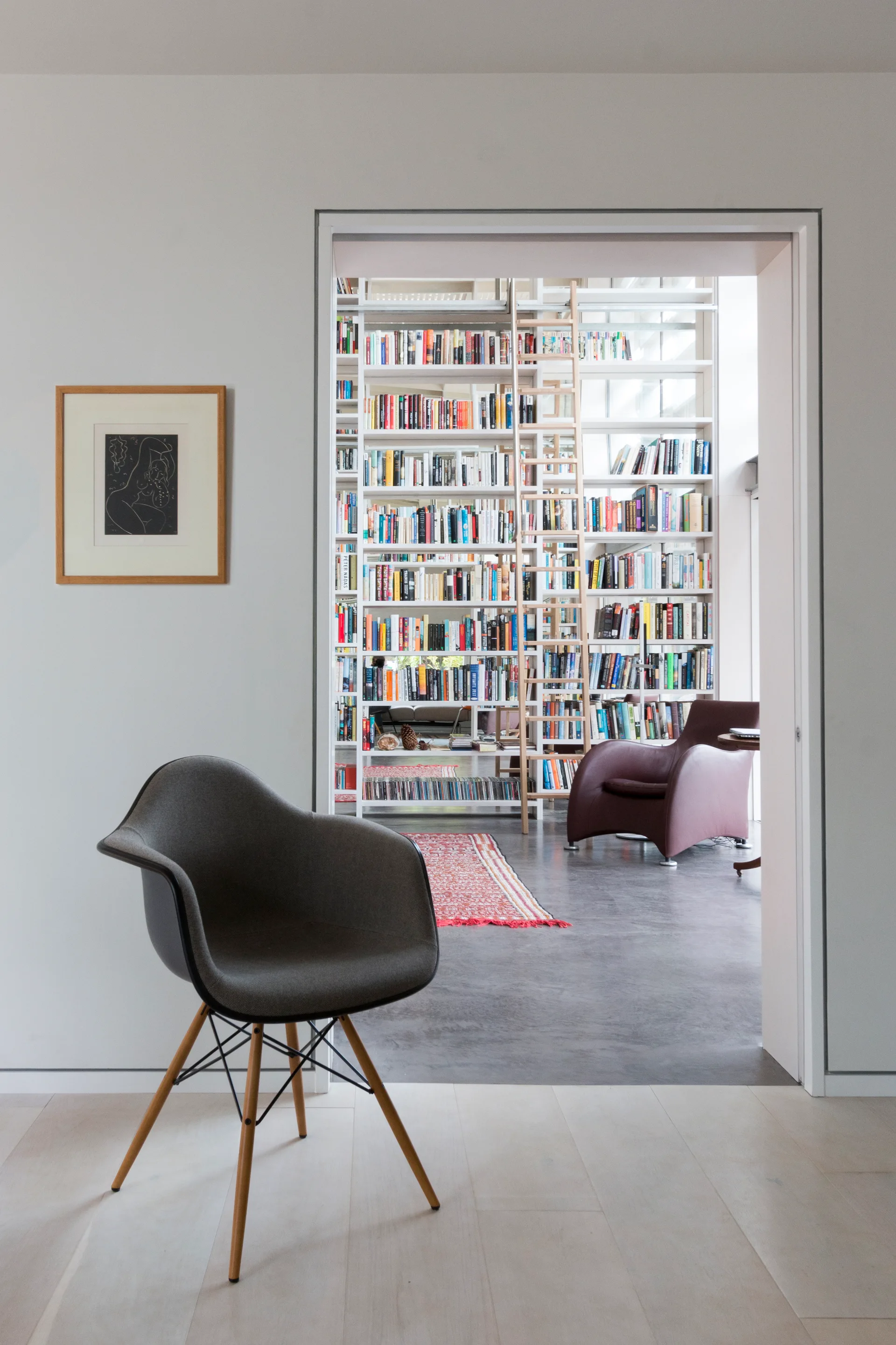 A view through a white doorway shows a home library with floor-to-ceiling white bookshelves, a wooden ladder, a patterned rug, and a modern chair.