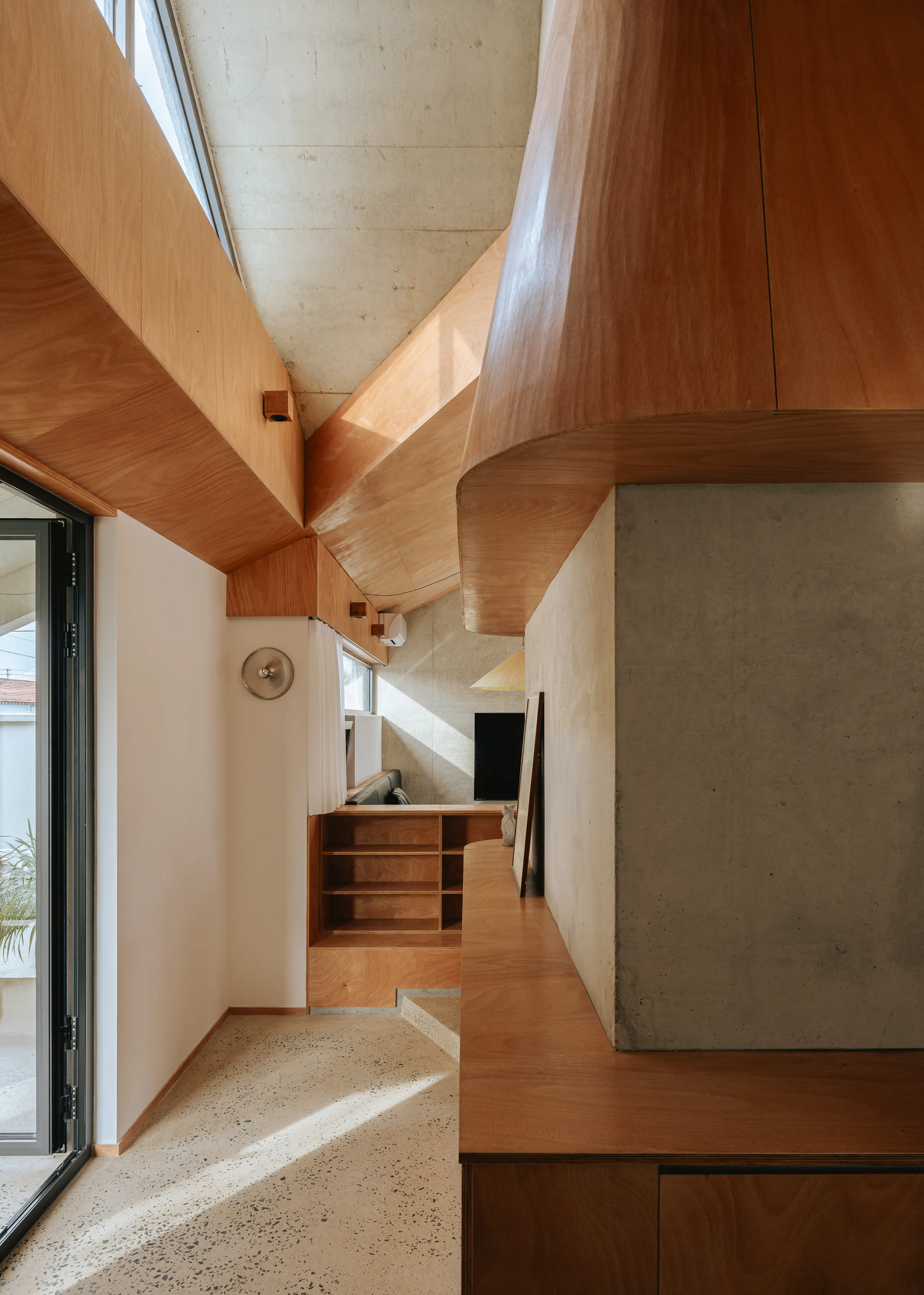 A modern residential interior featuring warm plywood built-in cabinetry, an angled concrete ceiling, and a light speckled terrazzo floor.