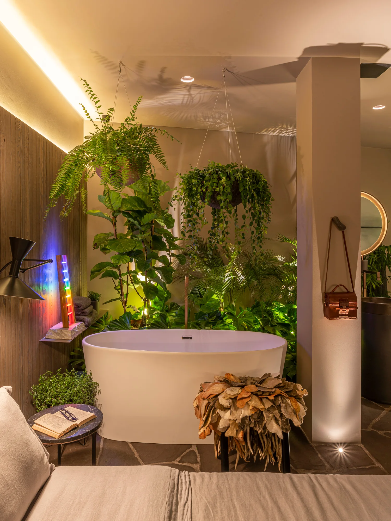 A white freestanding oval bathtub is surrounded by lush green hanging plants, stone flagstone flooring, and warm accent lighting.