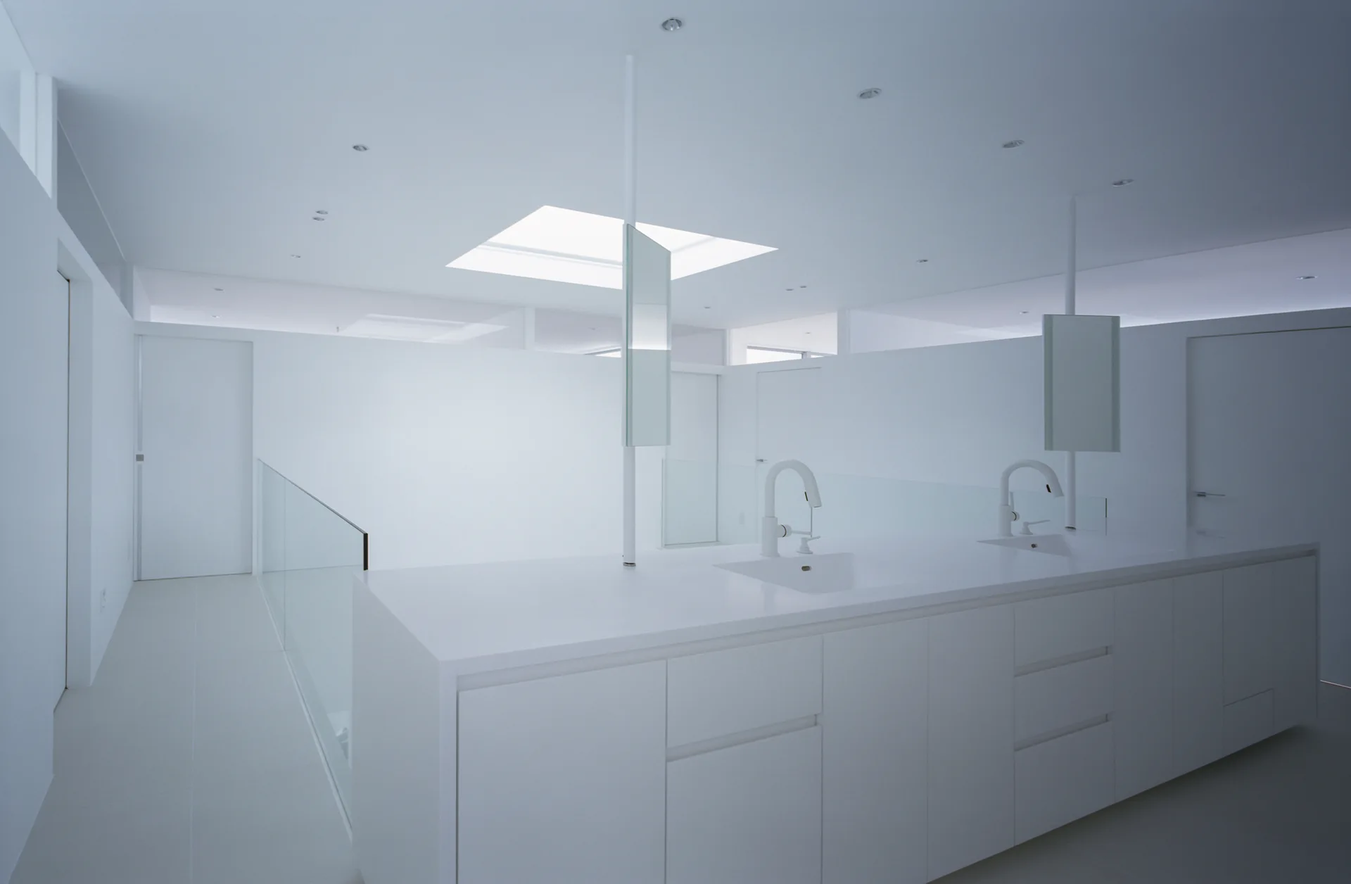 A minimalist all-white bathroom features a long double vanity with integrated sinks, white faucets, pole-mounted vertical mirrors, and a square skylight.