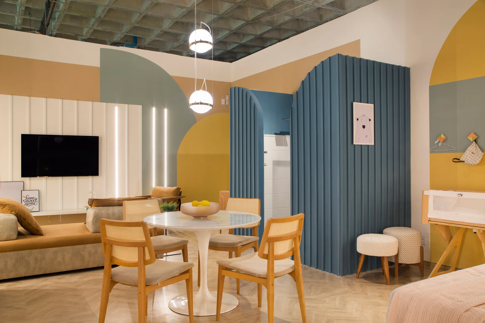 A modern living space features a round dining table with cane chairs, a blue ribbed partition wall, and colorful geometric wall paint.
