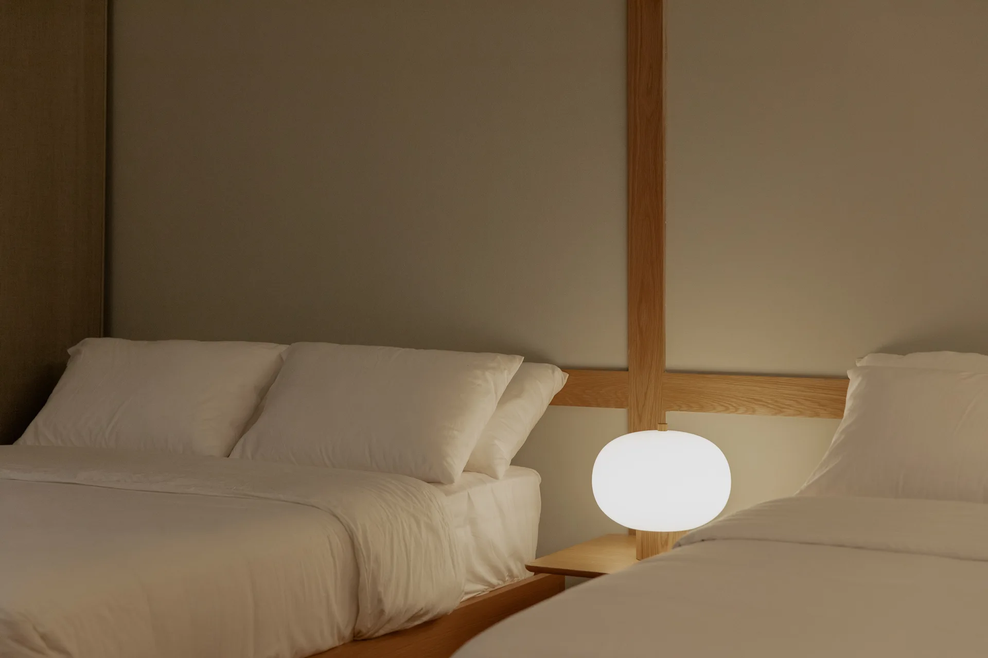 Two minimalist beds with white linens flank a low wooden nightstand with a glowing globe lamp against a beige wall with wood grid trim.