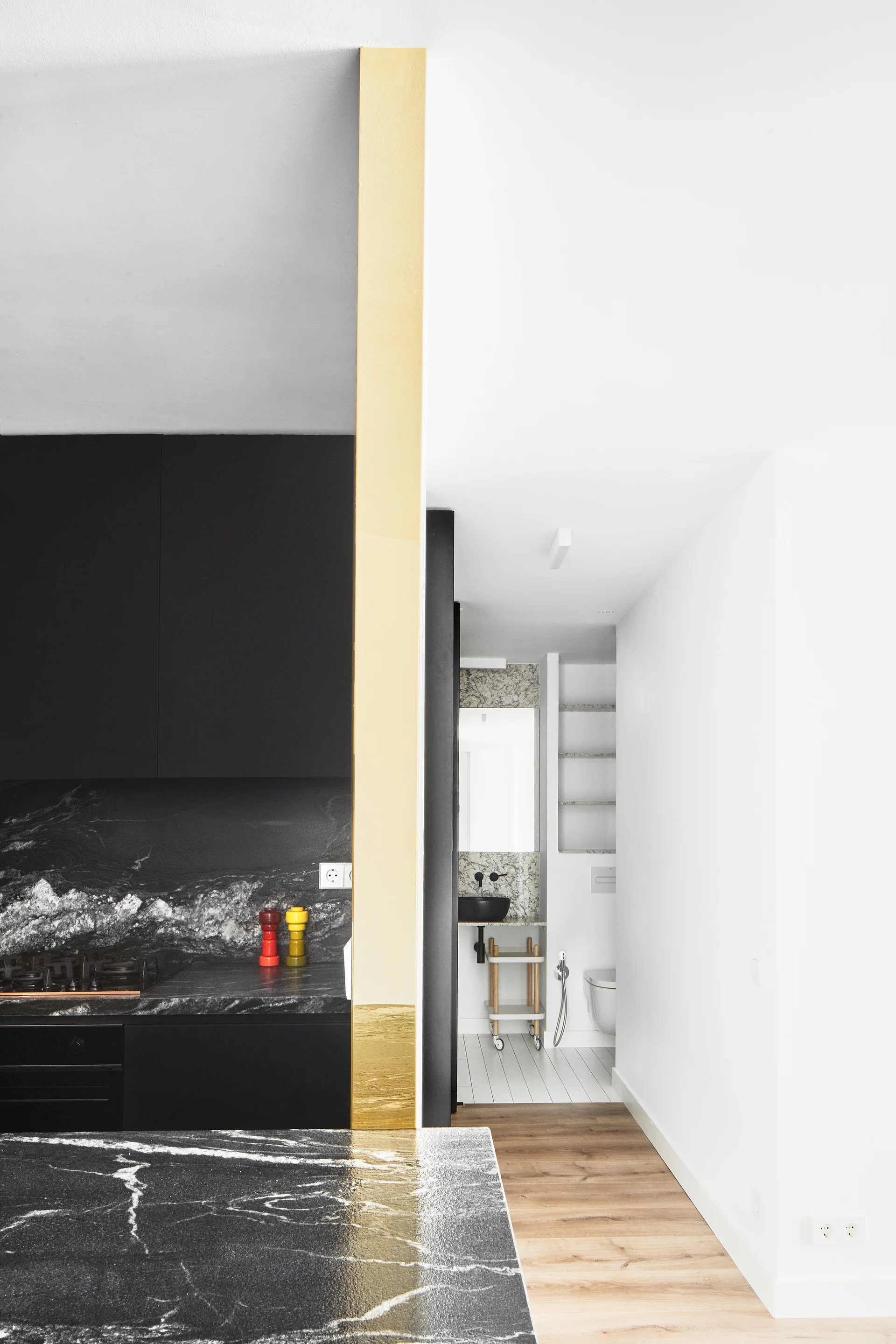A modern matte black kitchen with veined black marble countertops is separated by a gold-painted pillar from a white bathroom.