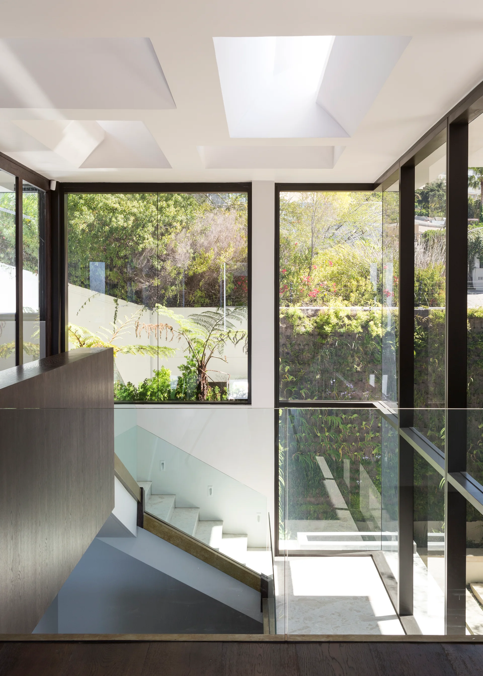 A modern interior staircase features light stone treads, glass railings, and recessed square skylights next to floor-to-ceiling glass windows.