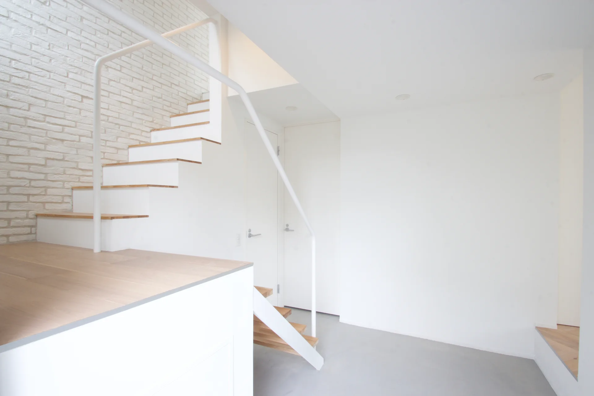 A minimalist white staircase features light wood treads and a metal handrail alongside a white painted brick accent wall.