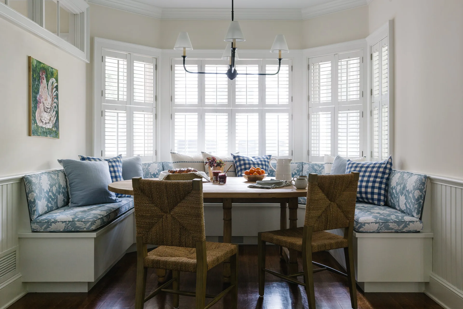 A breakfast nook features a round wooden table, woven chairs, and a white built-in banquette with blue and white patterned cushions set in a bay window.