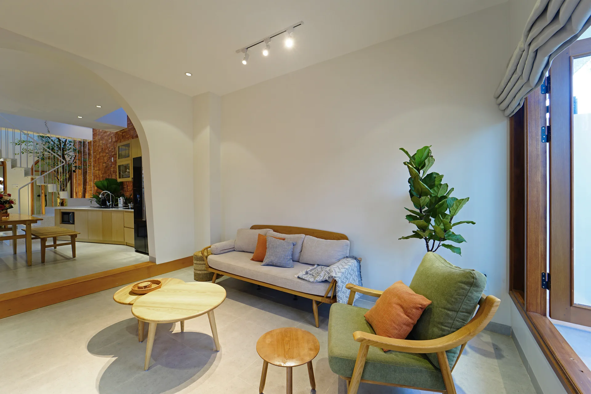 A bright living room features a light wood sofa, a green armchair, and a wide plaster archway looking into an adjacent kitchen.