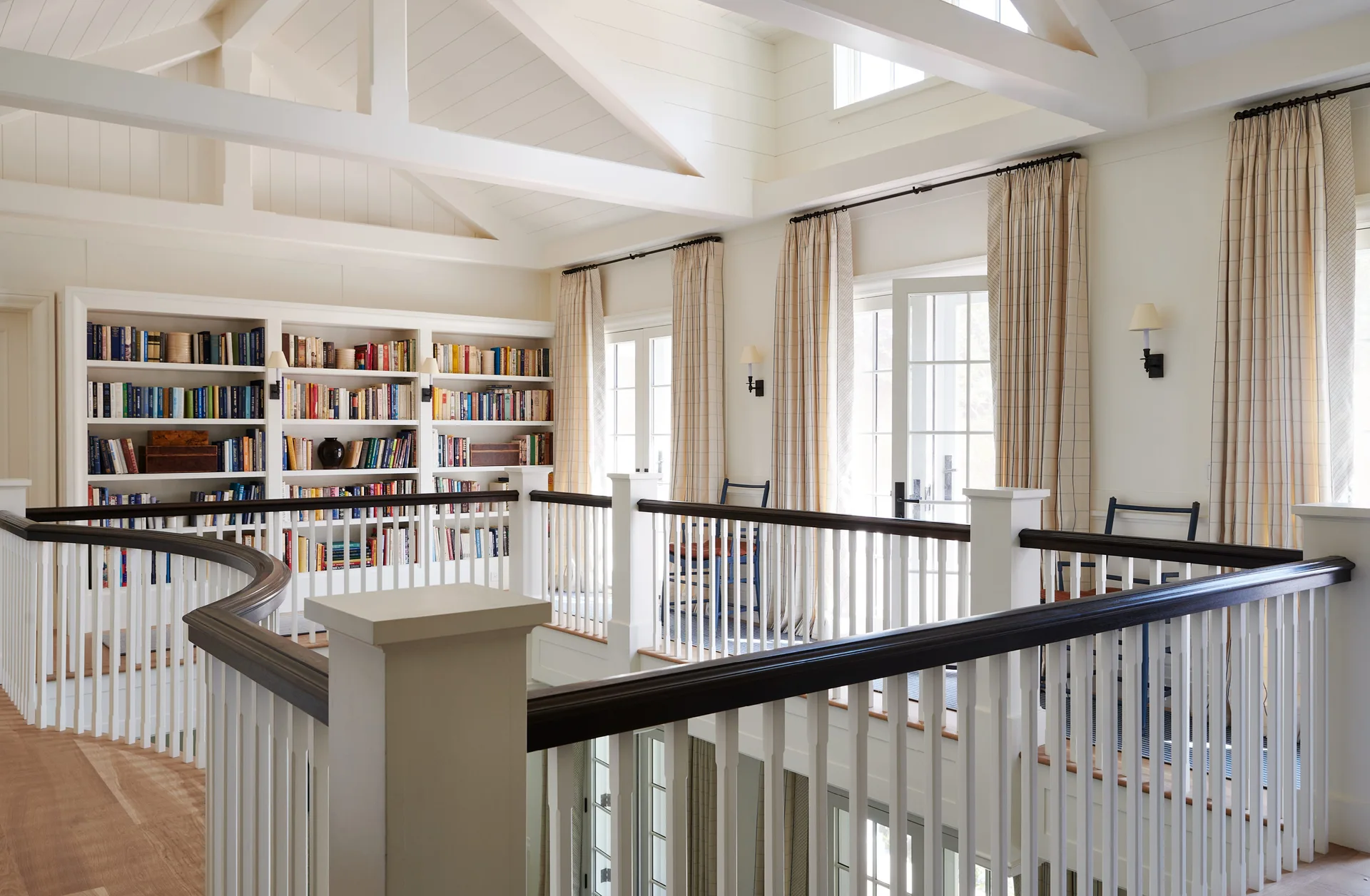 An upstairs landing features a curved white railing with a dark handrail, built-in bookshelves, and French doors under a beamed vaulted ceiling.