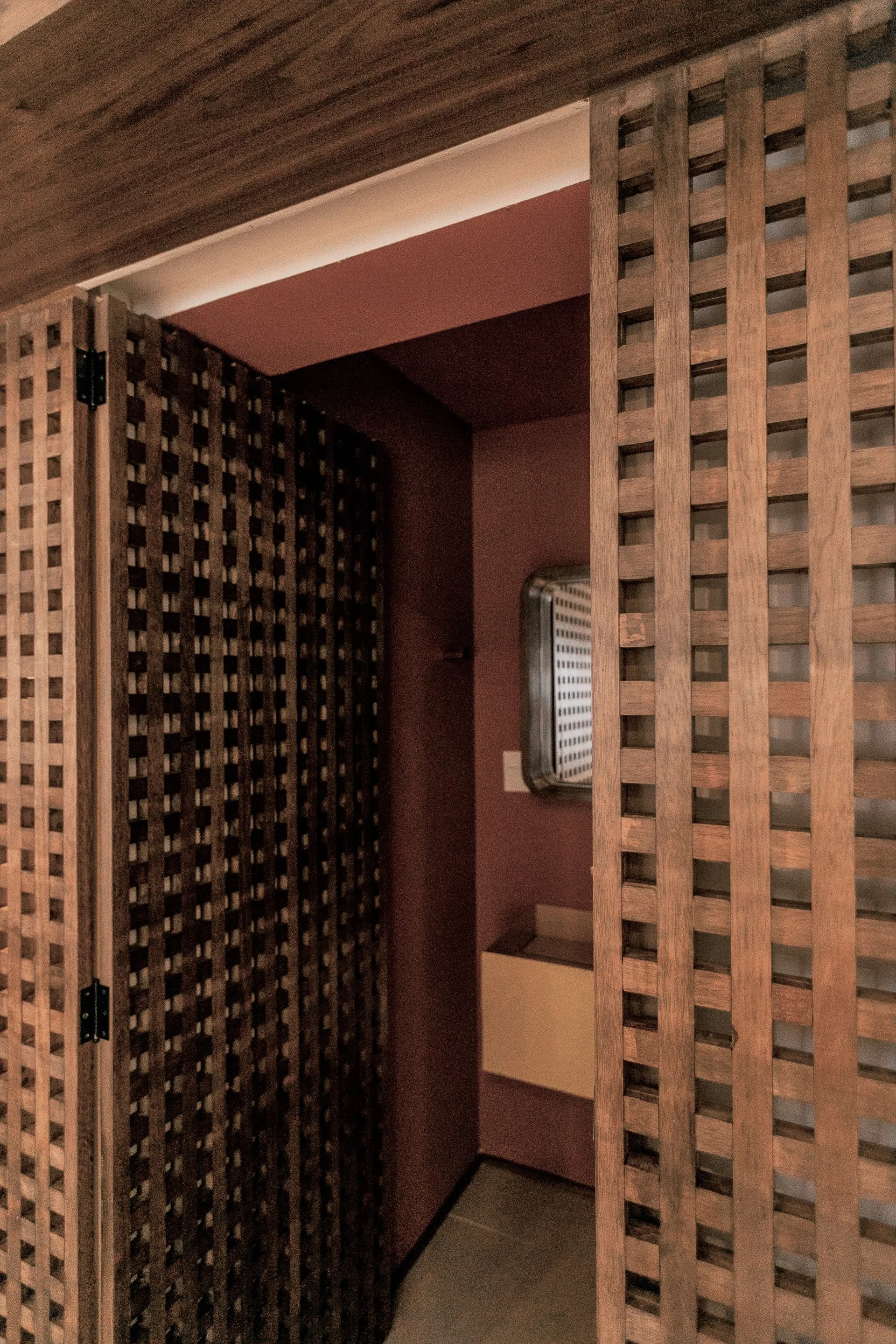 A wooden grid lattice screen partially conceals a dimly lit bathroom with dark red walls, a simple vanity, and a wall mirror.