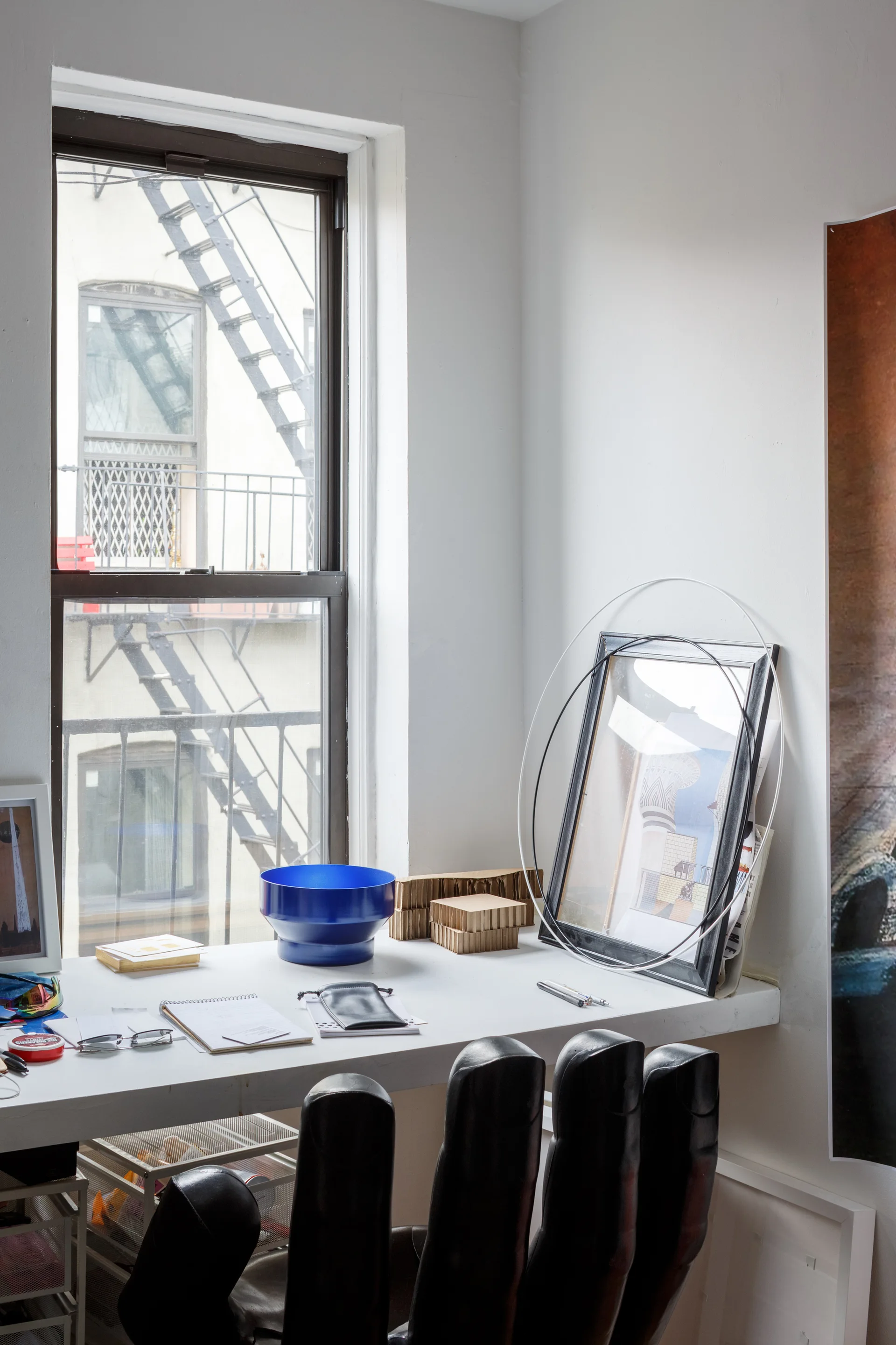 A white desk sits by a tall window showing a fire escape, holding a blue bowl, leaning art, and a black hand-shaped chair.