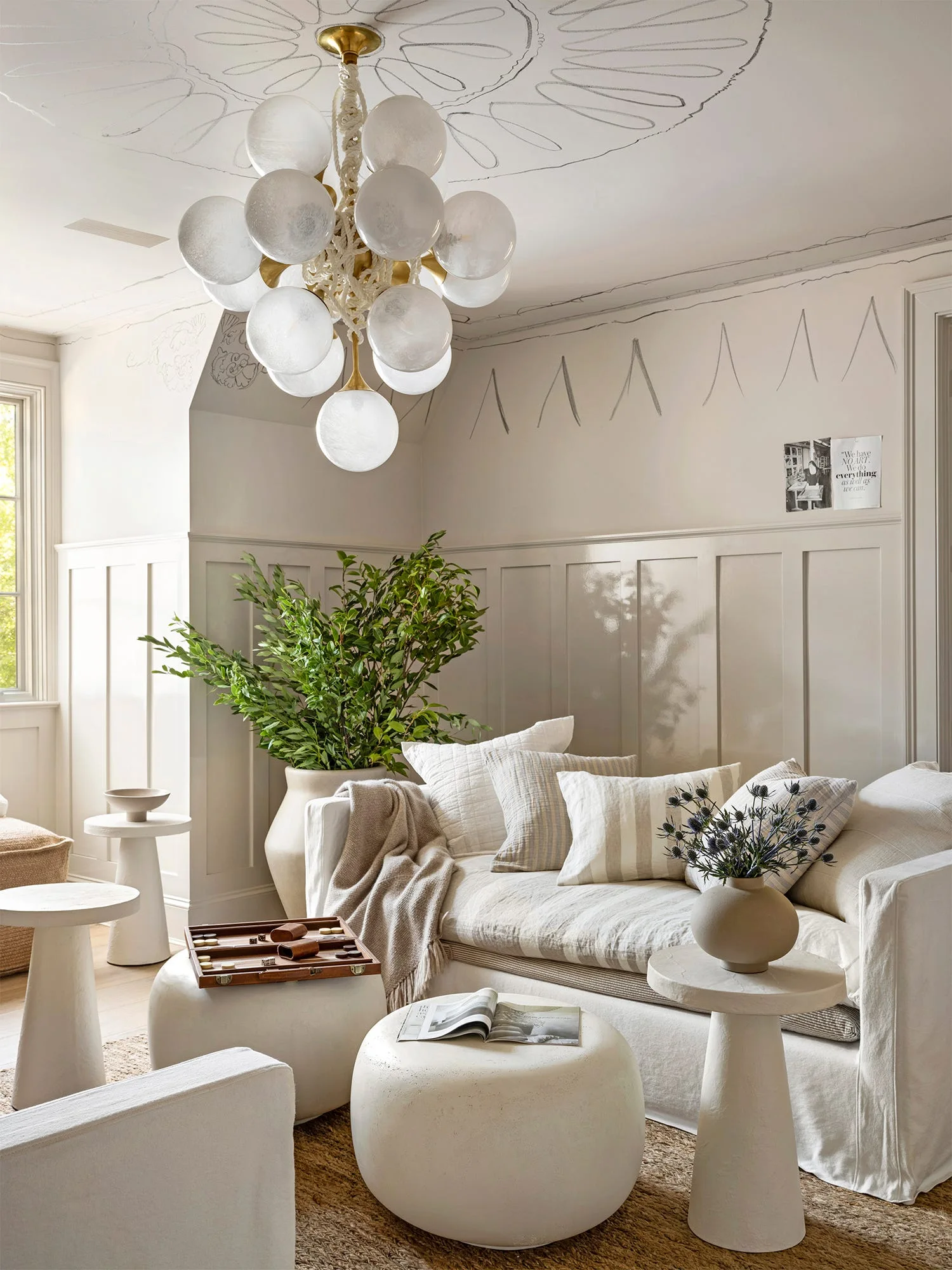 A cream linen sofa sits below off-white wainscoting decorated with hand-sketched charcoal lines and a multi-globe bubble chandelier.