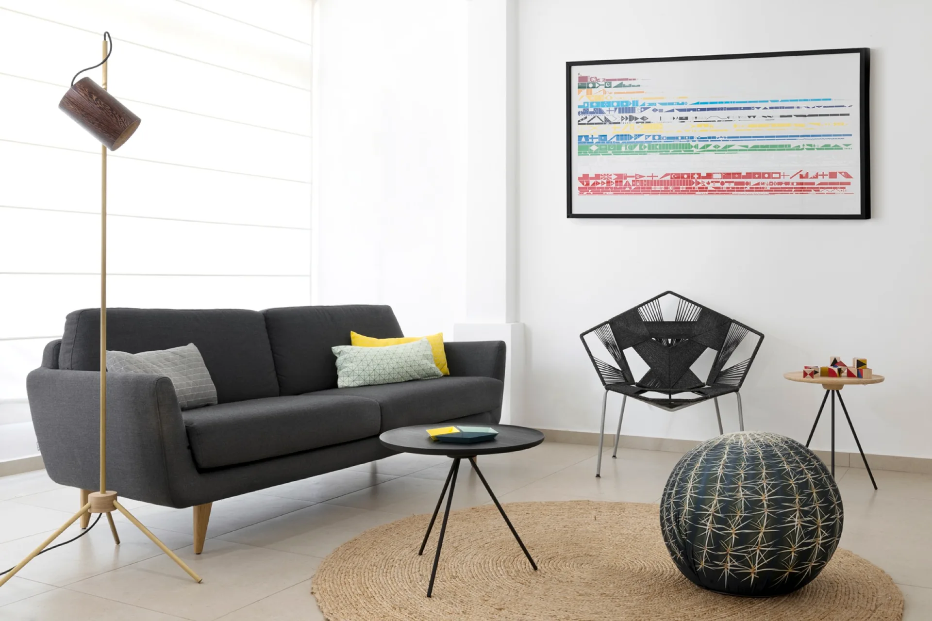 A bright modern living room featuring a charcoal grey sofa, a black geometric accent chair, and a round woven jute rug on tiled flooring.