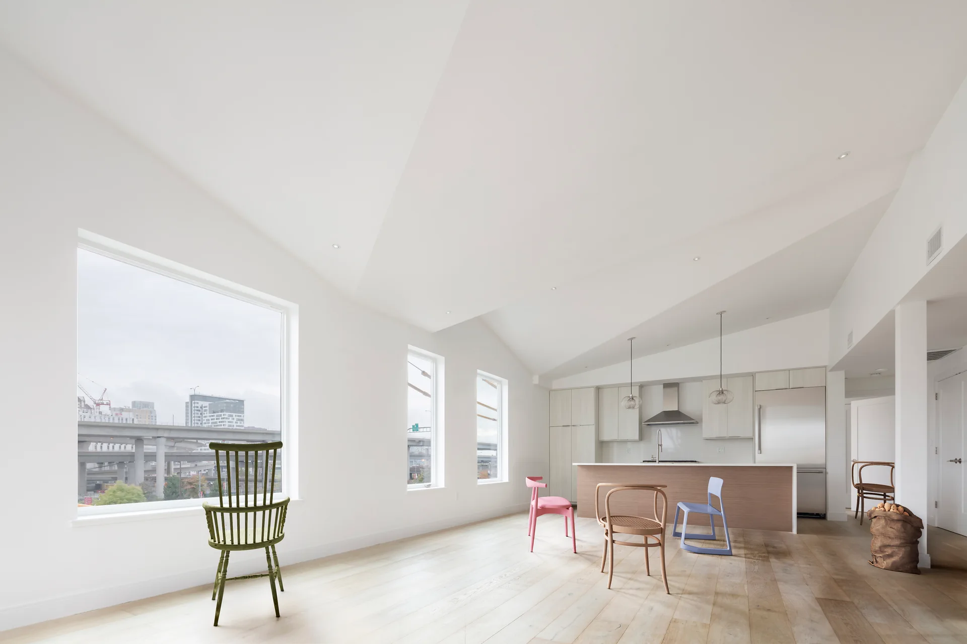 A spacious minimalist kitchen and dining area features white walls, sloped ceilings, light wood floors, and several mismatched colorful chairs.