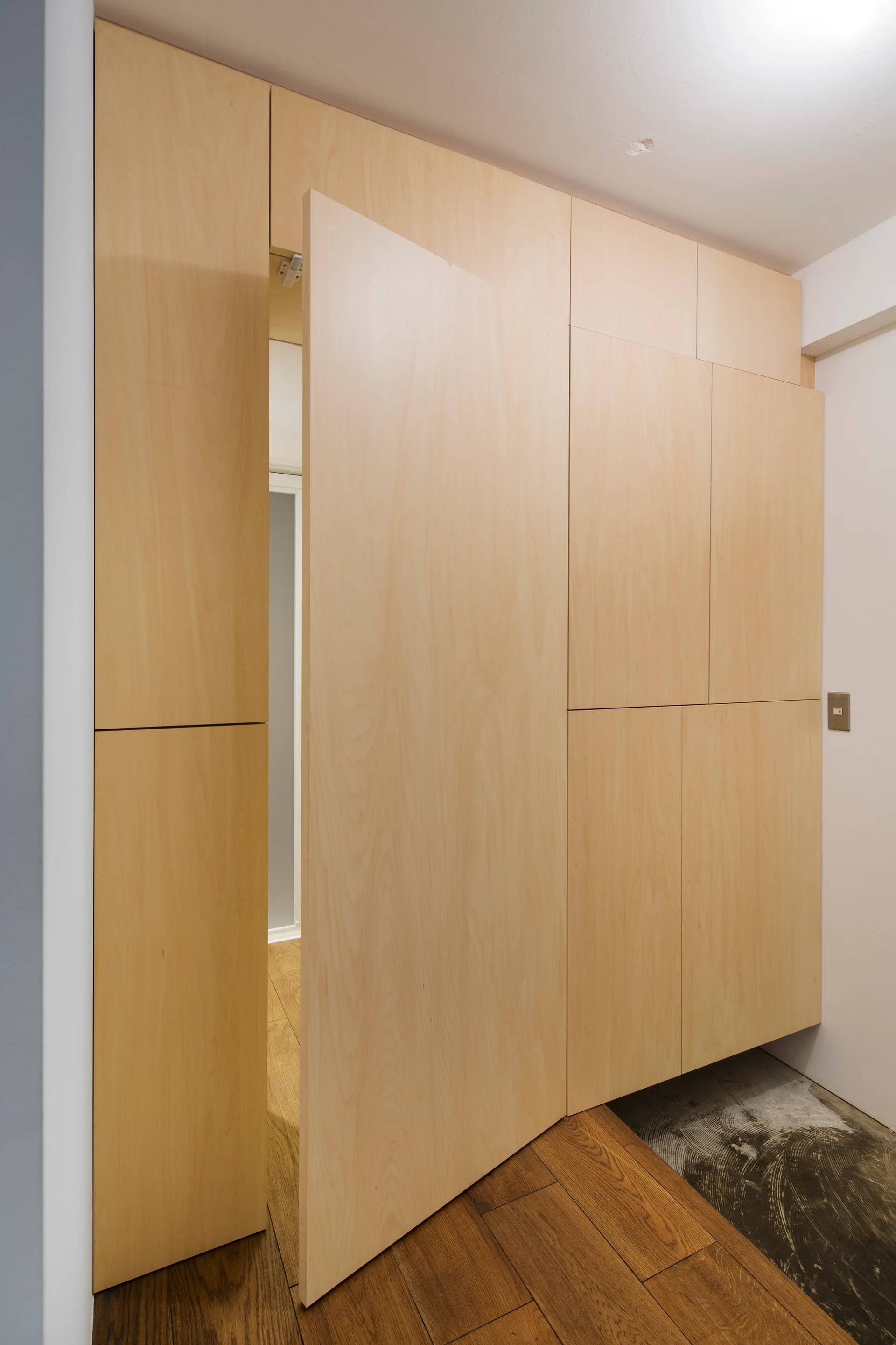 A minimalist light wood cabinet wall features a seamless pivoting door that opens into an adjacent room in a hallway.