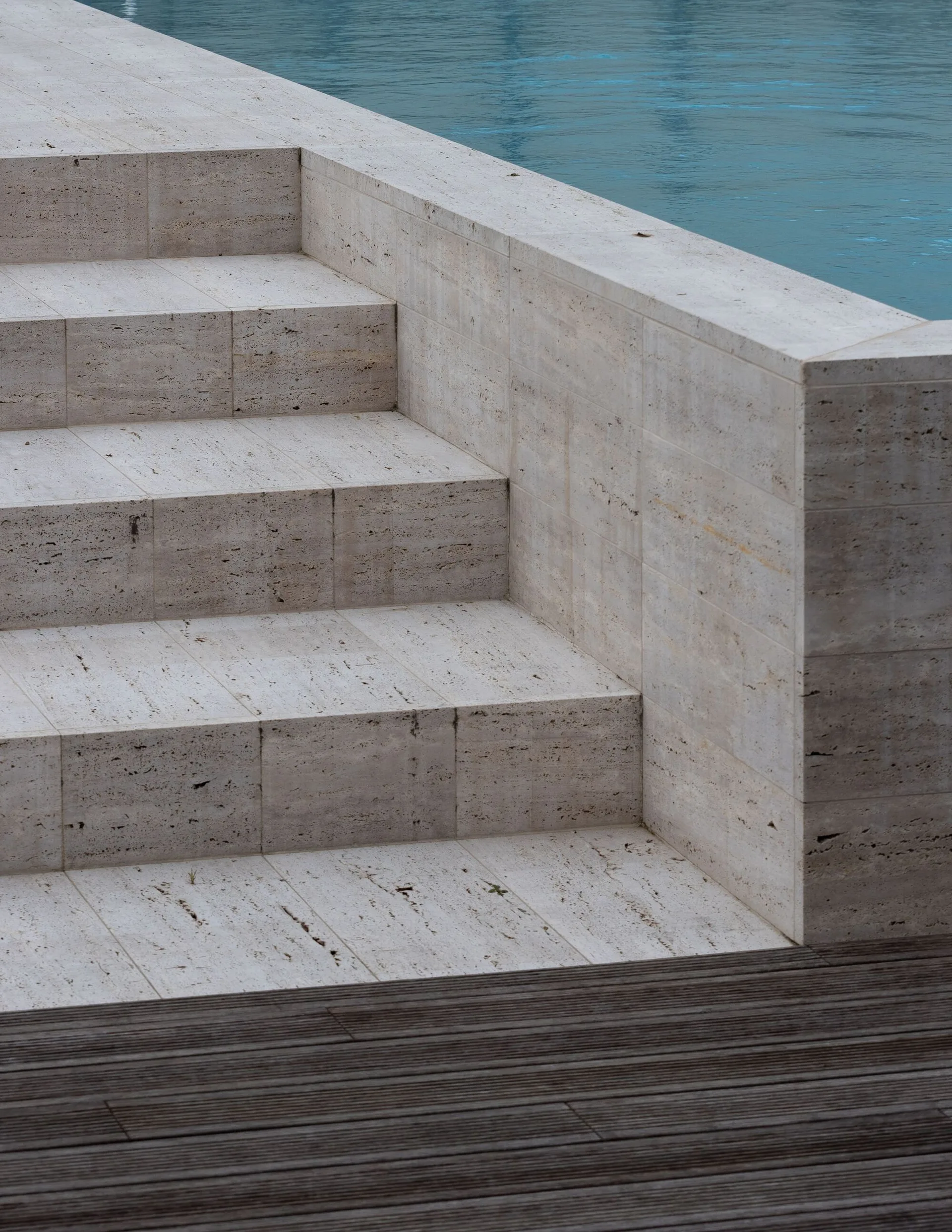 Beige travertine stone steps descend into a blue swimming pool next to a dark wood deck.
