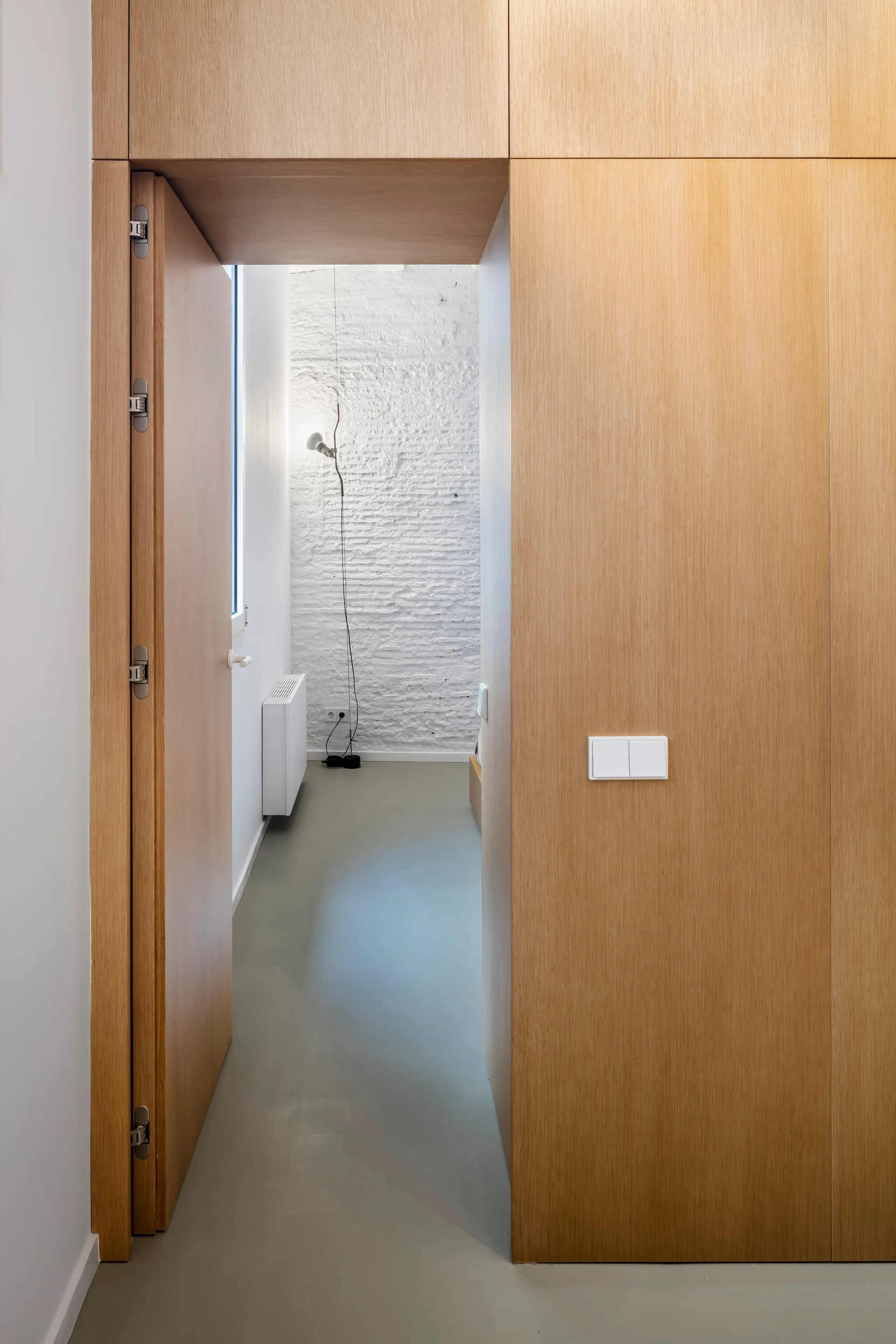 A view through a light wood paneled doorway into a minimalist bedroom featuring a textured white brick wall, grey flooring, and a slim floor lamp.