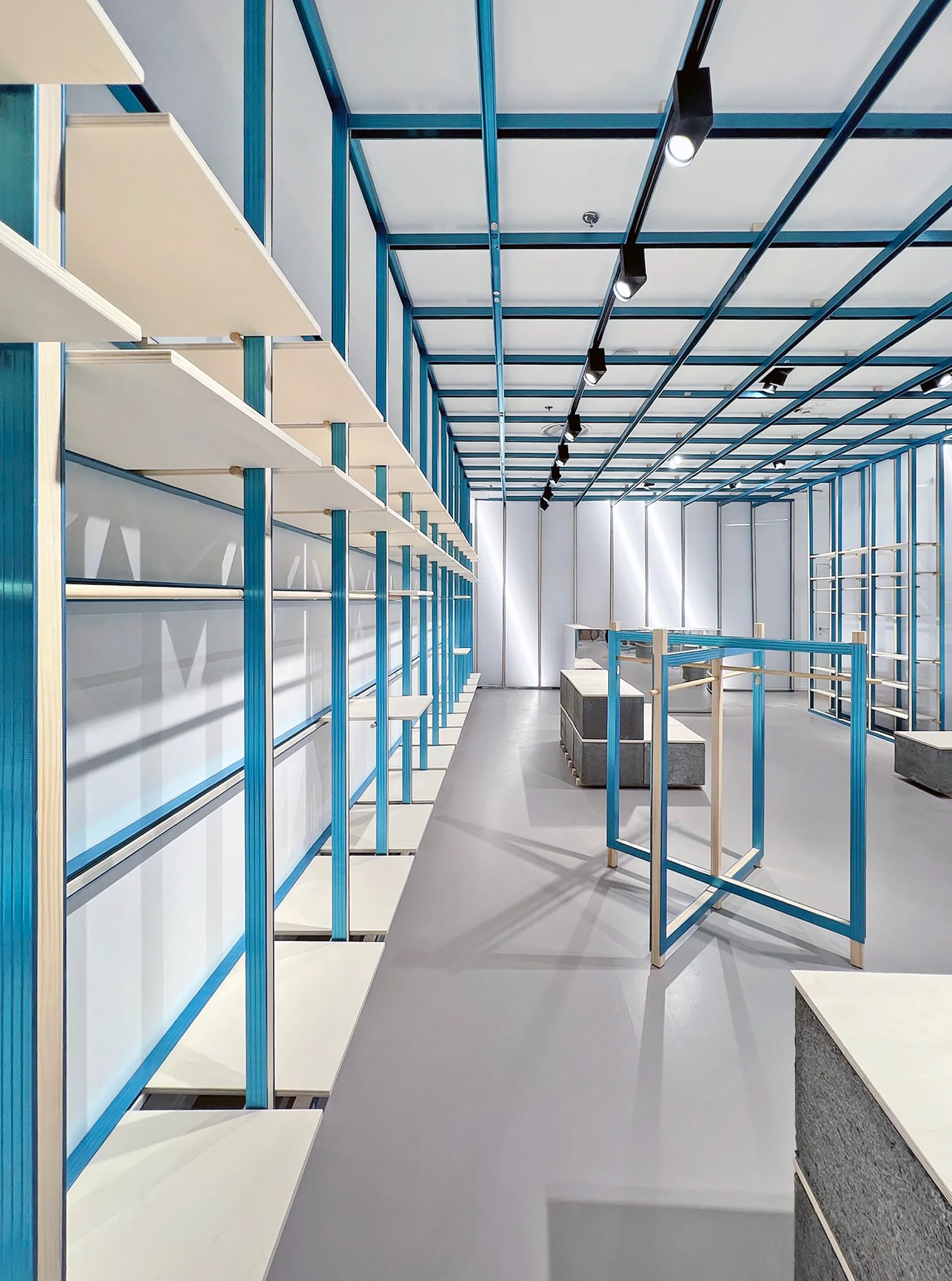 A modern minimalist commercial interior features a bright blue metal grid structure on the ceiling and walls with modular wood shelving.