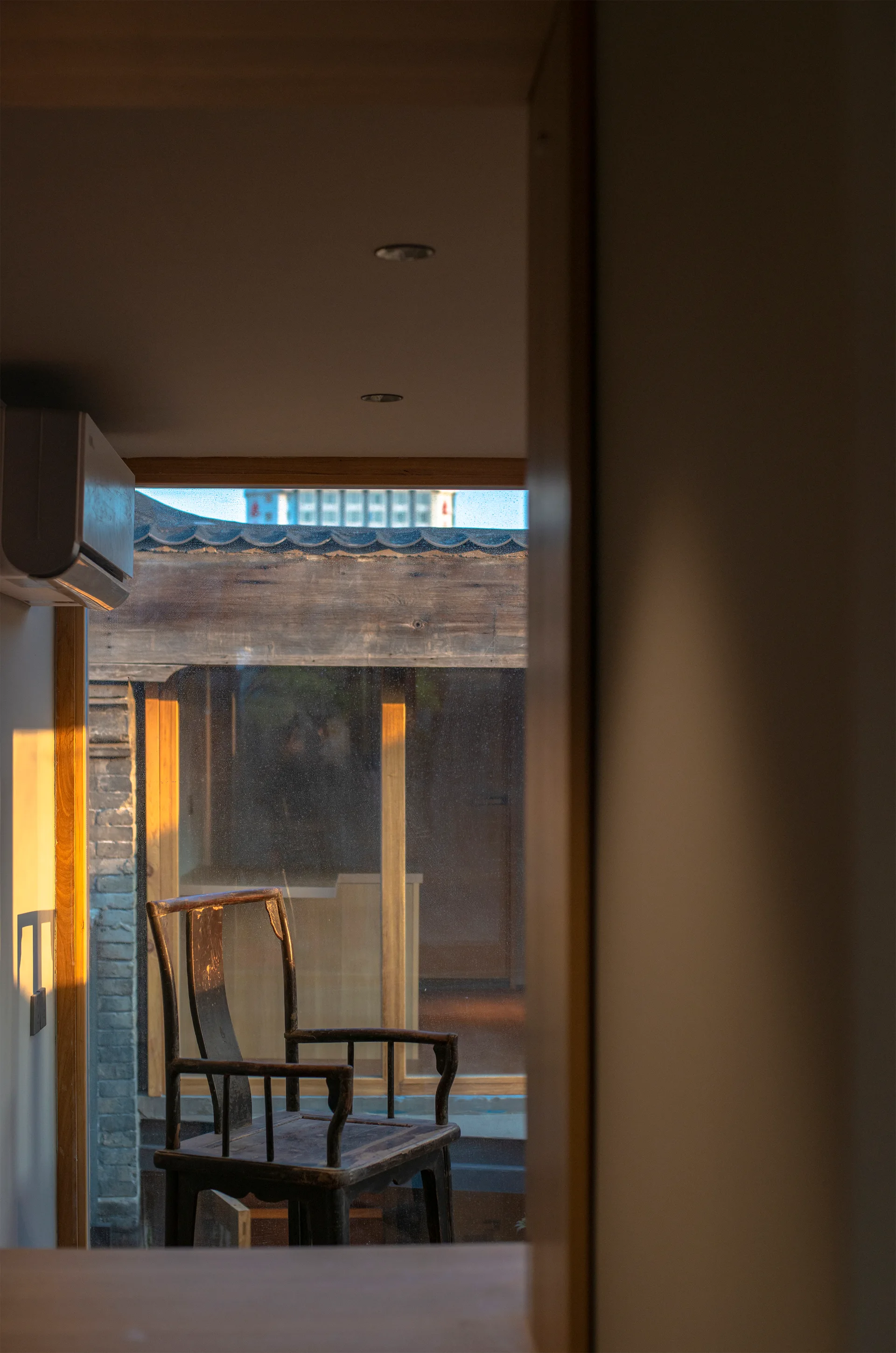 A dark wood traditional armchair sits in front of a window showcasing a curved tile roof structure under warm sunlight.