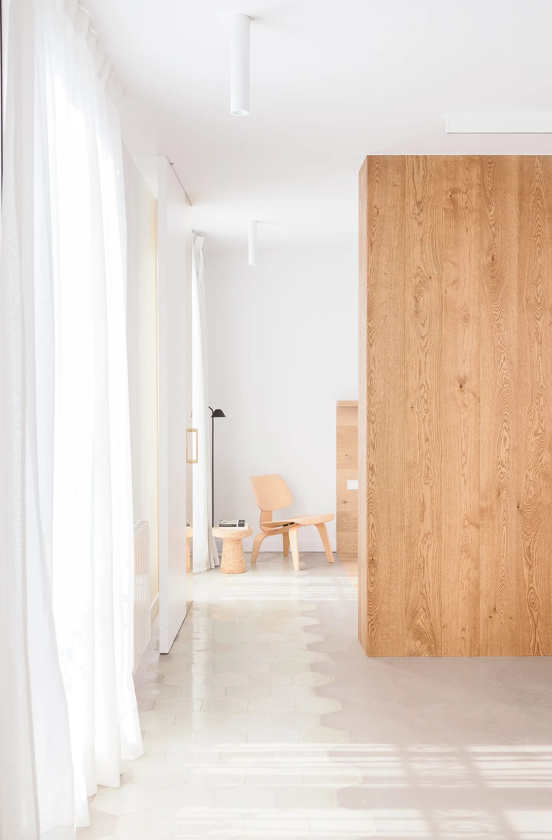 A bright, minimalist hallway features a tall wood-grained partition wall, white sheer curtains, and light-toned hexagonal floor tiles leading to a wooden chair.