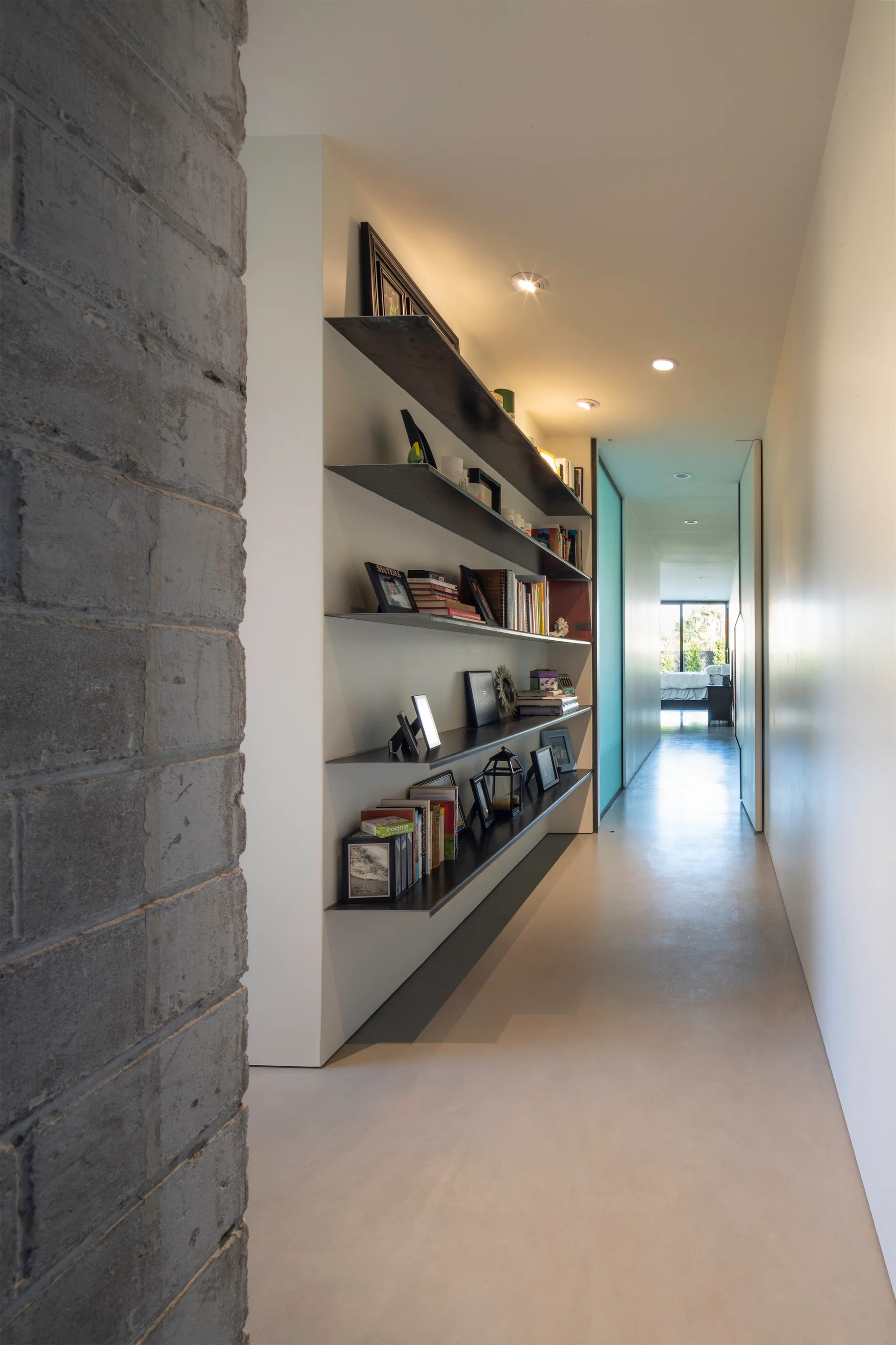 A long minimalist hallway features recessed ceiling lights, a textured gray brick wall, and floating dark metal shelves displaying books and frames.