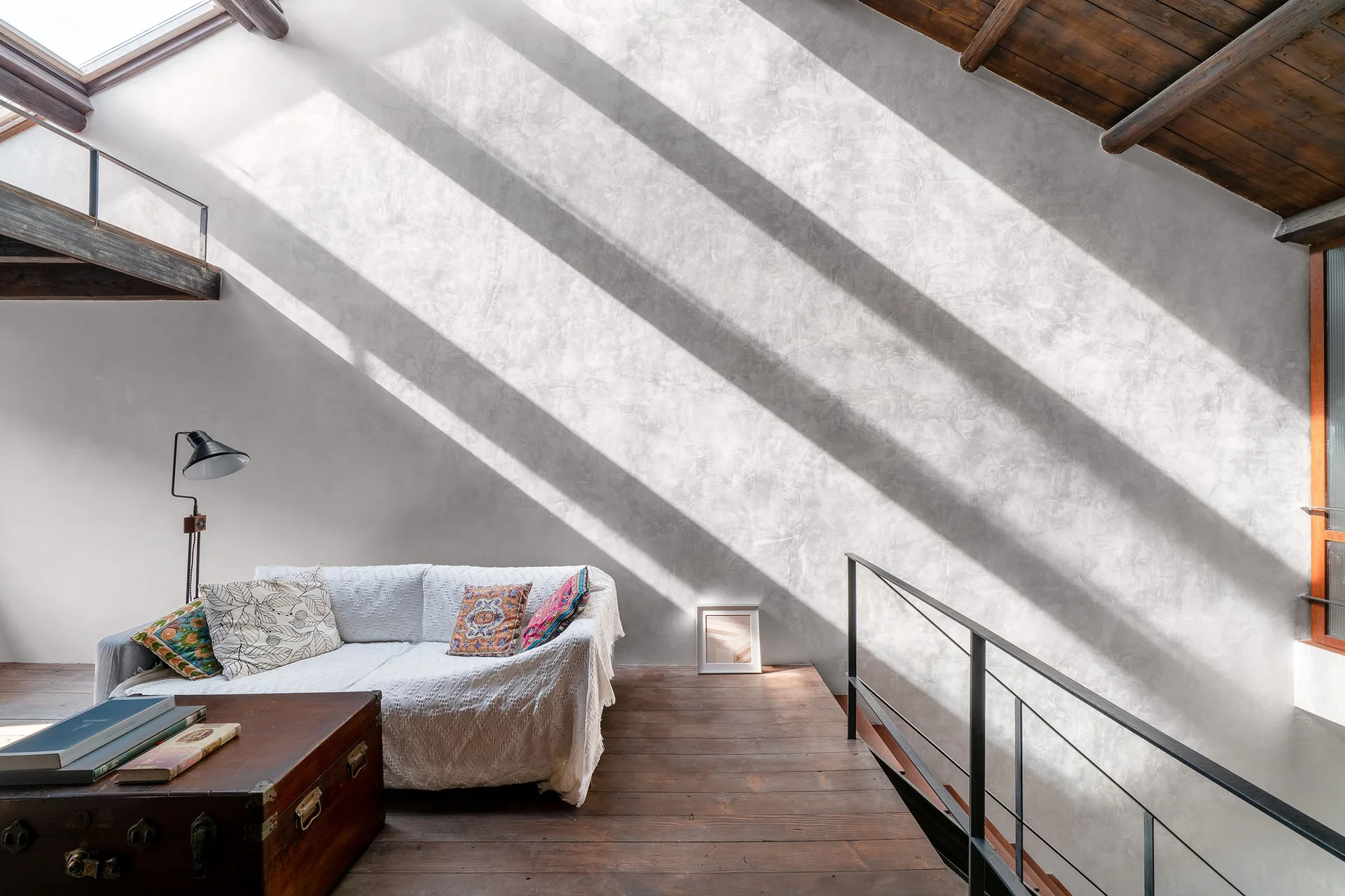 A sunlit loft sitting area features a white sofa, a vintage wooden trunk table, and a sloped ceiling with exposed beams and a skylight.