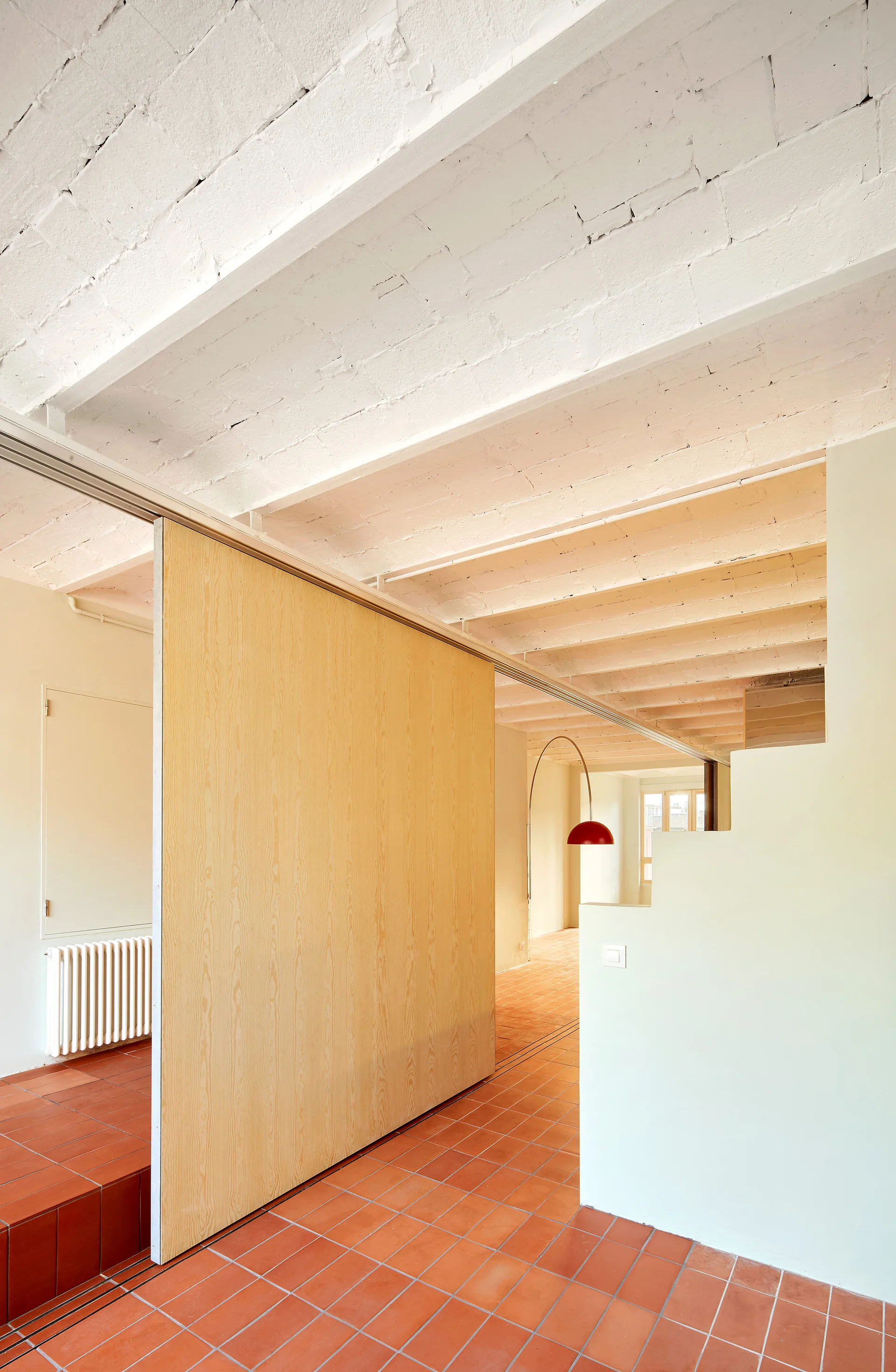 A large light wood sliding partition stands on a terracotta tiled floor beneath painted white exposed ceiling beams in a minimalist interior.