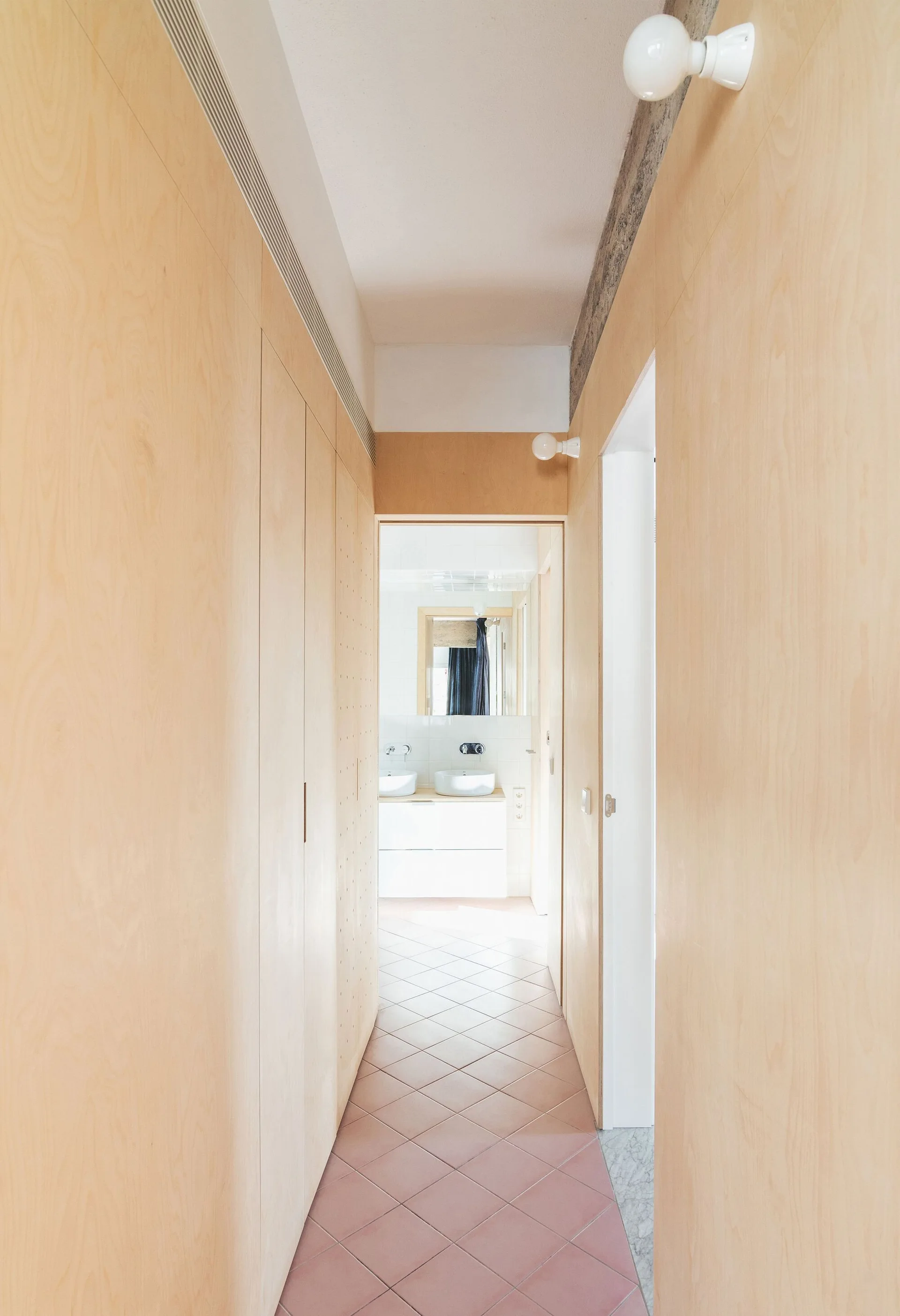 A narrow hallway featuring light wood paneled walls, diagonal terracotta floor tiles, and simple wall-mounted bulb lights leading to a white bathroom.