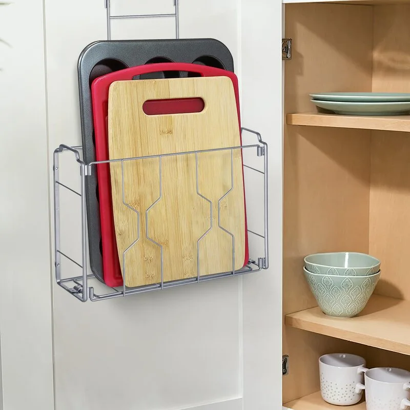 A silver metal wire rack hangs over a white cabinet door, holding wooden and red cutting boards next to an open cupboard with shelves.