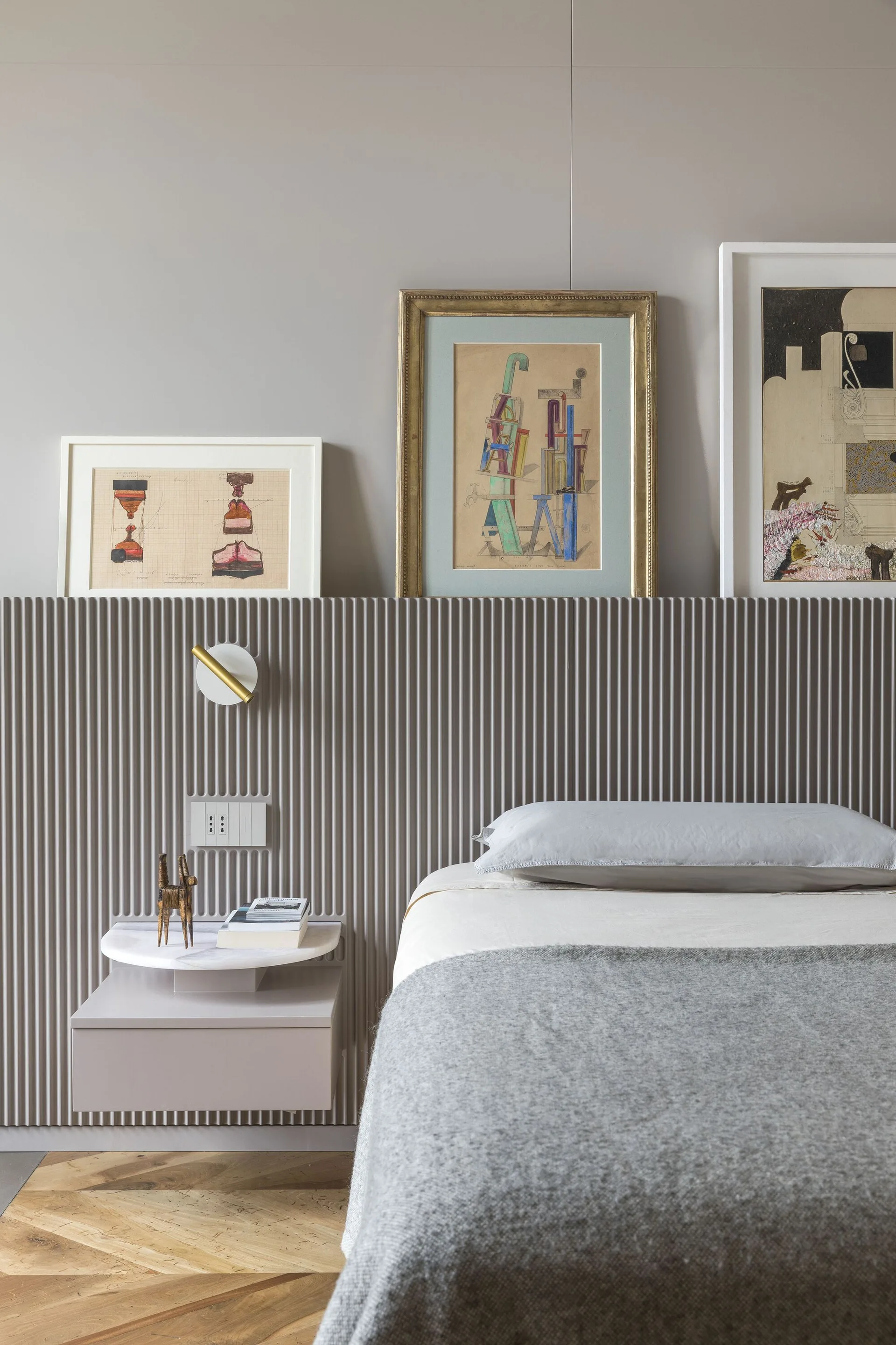 A bedroom features a taupe fluted headboard panel with framed artwork resting on its ledge, a floating nightstand, and herringbone flooring.