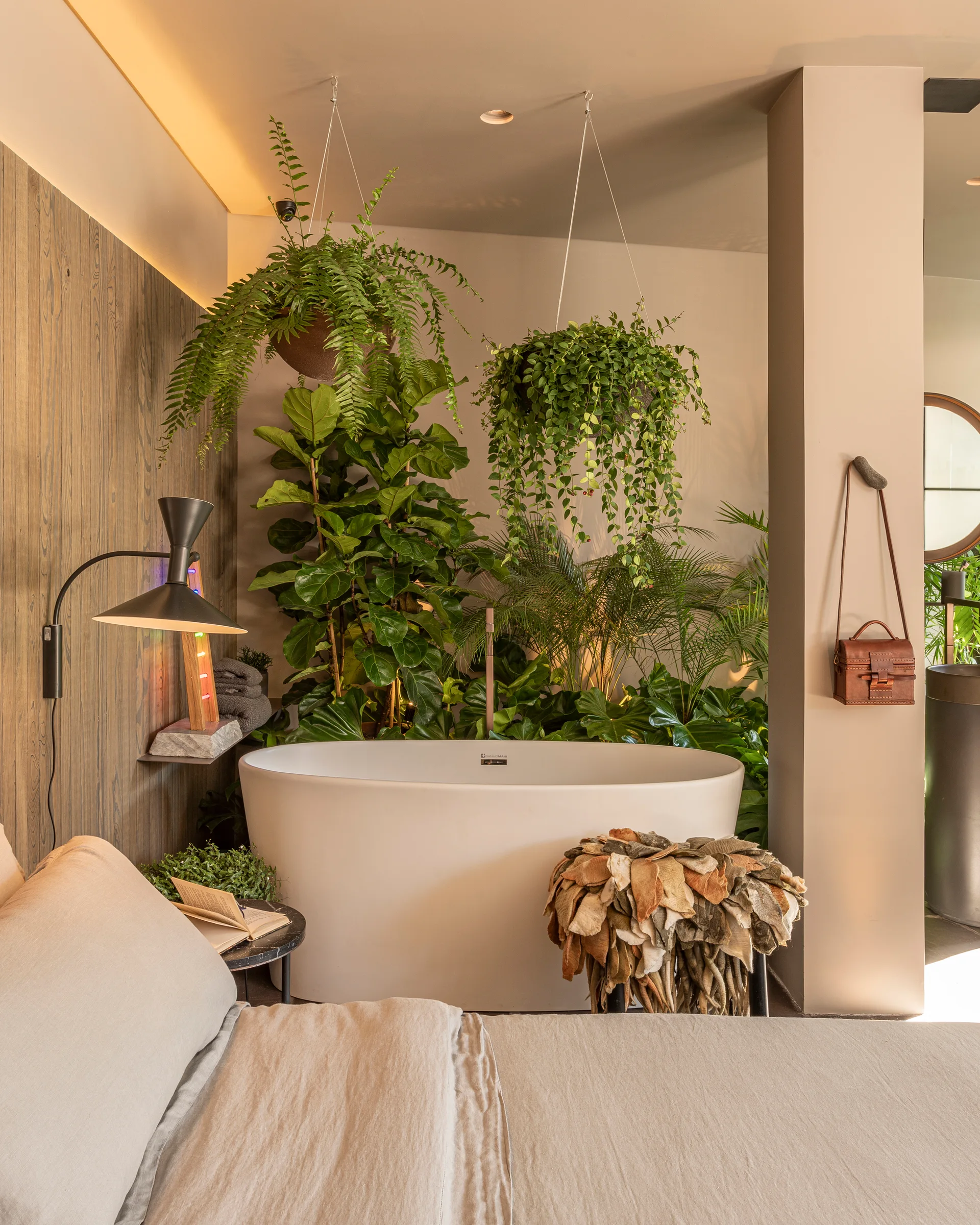 A modern bedroom features a white freestanding bathtub surrounded by dense green houseplants, wood wall paneling, and a bed in the foreground.