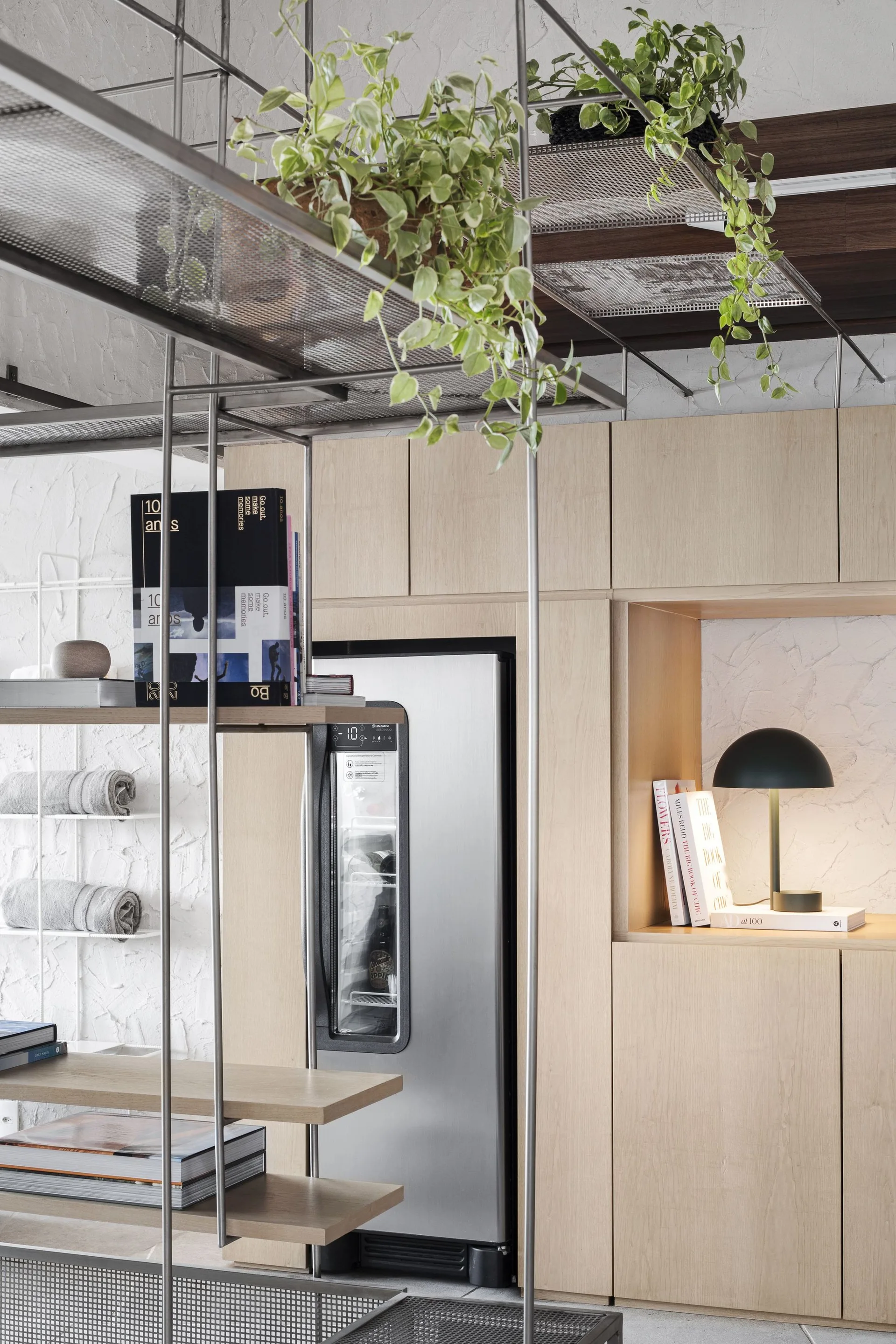 Modern light wood kitchen cabinetry features an integrated wine cooler alongside a metal grid shelving unit with green plants and books.