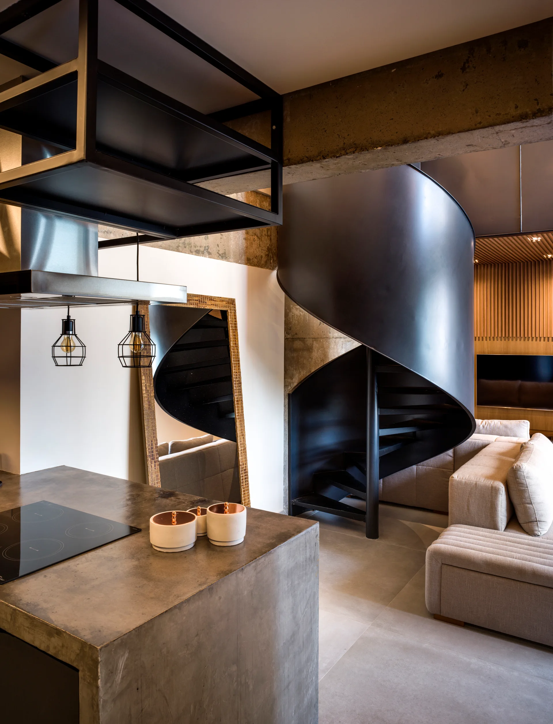 A modern open-plan loft featuring a concrete kitchen island, a black metal spiral staircase, and a beige sectional sofa.