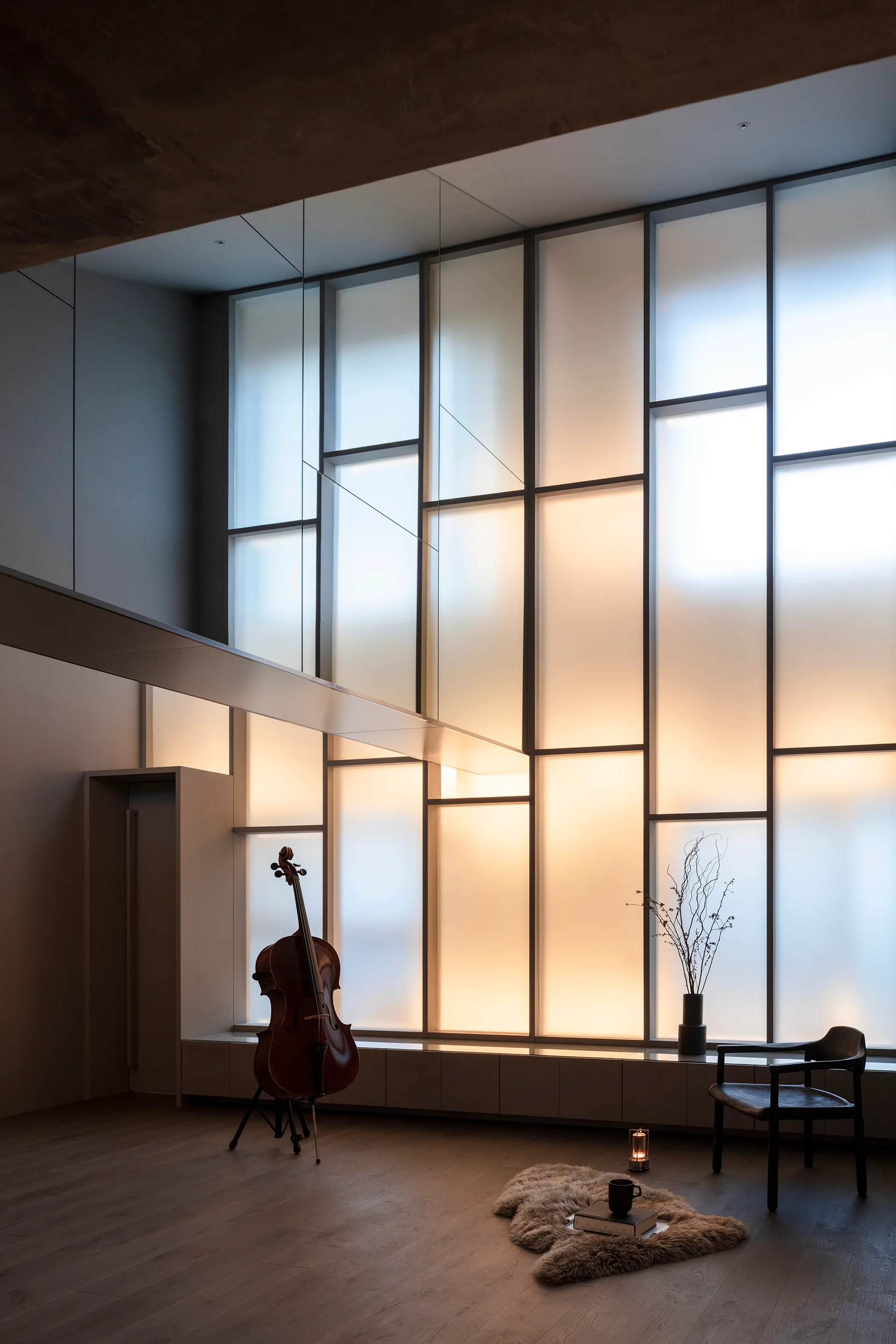 A minimalist room featuring a large backlit frosted glass grid window, a wooden cello on a stand, and a dark accent chair on wide plank floors.
